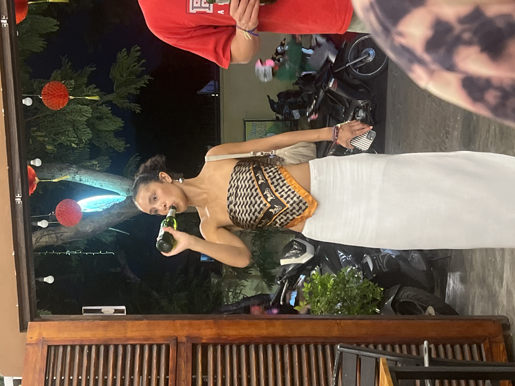 The image shows a nighttime scene in Đà Nẵng, Vietnam. The main subject is a young woman with dark hair, wearing a patterned scarf top and a white skirt. She is holding a green glass bottle to her mouth, appearing to drink from it. Her left arm is bent at the elbow, and her right arm is extended. A beige purse is slung over her shoulder.
In the background, there are several motorcycles parked. Red paper lanterns and string lights are visible, suggesting a festive or decorative setting. The lighting indicates it's nighttime, with the bright lights from the lanterns and possibly artificial lights illuminating the scene. The backdrop appears to be an outdoor area, possibly a street or market, with some greenery and trees.
To the left of the woman, another person is partially visible, wearing a red t-shirt. A part of a person's arm with a patterned sleeve is visible on the right side of the frame. The overall mood seems casual and relaxed, consistent with an evening outing. There is no discernible weather information. The presence of motorcycles and the specific lanterns could be indicative of a Southeast Asian location, further supported by the given location context. No text is clearly visible or legible in the image.