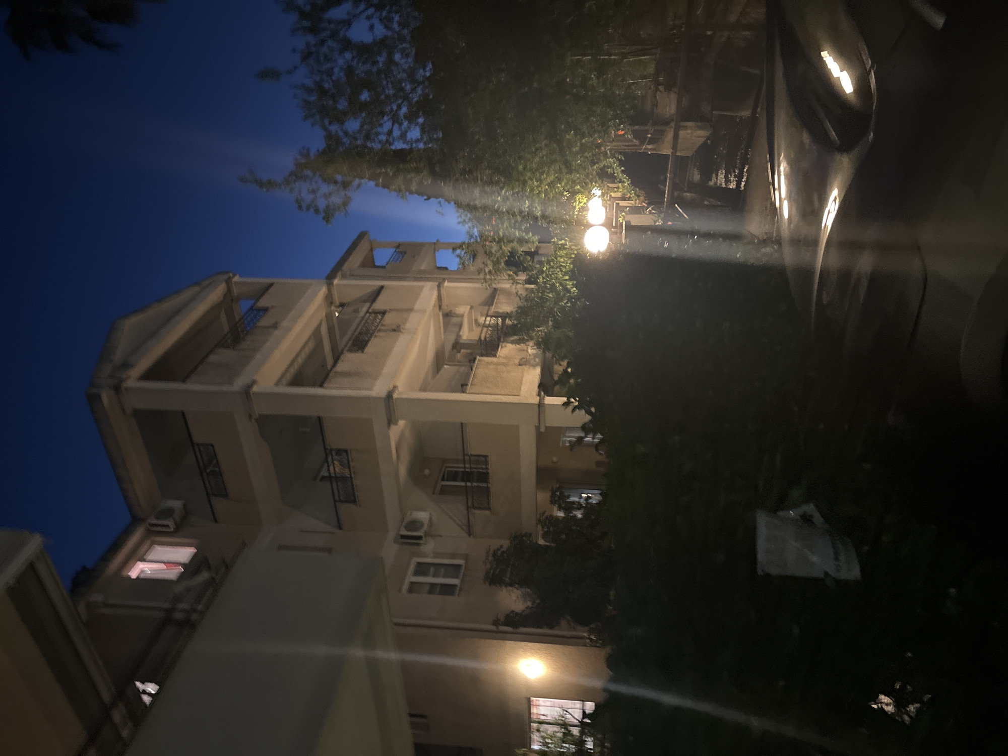 The image shows a building at night, with streetlights and illuminated windows. The building appears to be multi-story with balconies and a partially constructed upper floor. Trees and foliage are visible around the building, and a car is parked to the right, its side reflecting the ambient light. The sky is dark blue, indicating it is nighttime. A white plastic bag is visible on the ground amidst the foliage. The scene suggests an urban or semi-urban environment, possibly residential. There is no text visible in the image.