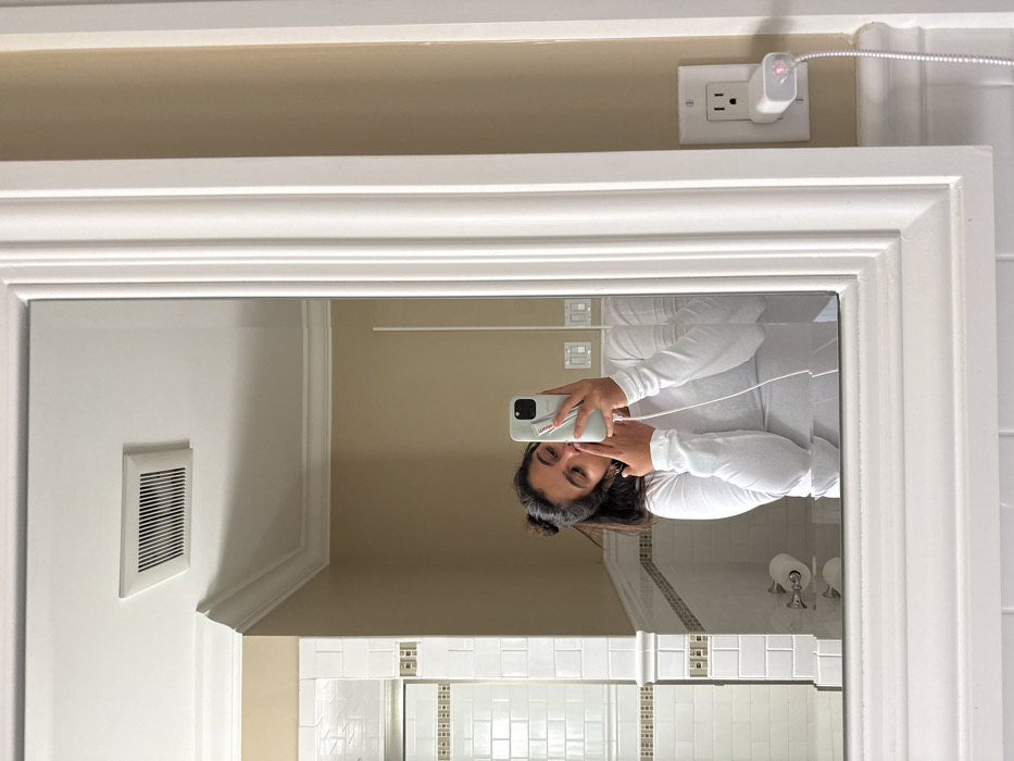 The image depicts a mirror selfie taken by a female subject in a private bathroom located in Princeton, United States. The subject, visible from the chest up in the mirror's reflection, has dark hair partially tied back and wears a white long-sleeved top. Her left index finger is positioned near her lips in a gestural pose. Her right hand holds a light-colored smartphone, which has a red/pink accessory attached to its back. A white braided charging cable extends from the phone to a white wall charger, which is plugged into a standard white duplex electrical outlet on the beige wall immediately adjacent to the mirror's white ornate frame. The charger emits a pink indicator light. The reflected bathroom interior features white subway tiles with a decorative brown and white accent border, beige painted walls, white crown molding, a rectangular white ceiling vent, and a white toilet paper holder with a roll.