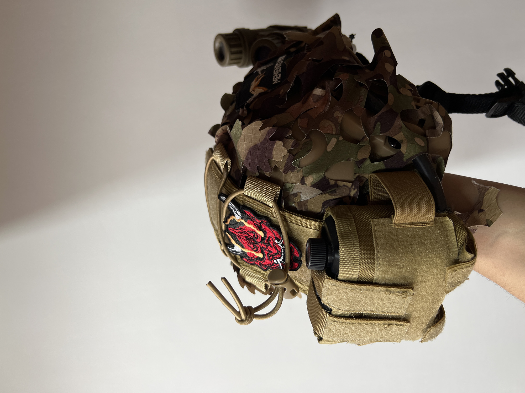 The image features a tactical helmet or headgear system, presented against a plain, light-colored background, suggesting an indoor setting. The headgear is largely covered by a camouflage fabric overlay, intricately cut into leaf-like shapes in a multi-cam pattern, providing a ghillie-style concealment effect.
On top of the helmet cover, a small rectangular patch is visible with the text "MEMENTO". A cylindrical optical device, possibly a night vision monocular or similar attachment, is mounted towards the front of the helmet.
Attached to the left side of the helmet (from the viewer's perspective) is a tan-colored utility pouch. A prominent red and black patch, depicting a stylized, fierce dragon or Oni mask-like face with white and black detailing, is affixed to this pouch. A tan bungee cord with a plastic toggle is also present on this side.
On the right side of the helmet, another tan-colored pouch is visible, secured with velcro straps, likely designed to hold accessories such as a battery pack or counterweight. A black strap system with a buckle is also seen towards the rear of the helmet, indicating part of its suspension or retention system.
A light-skinned human arm, showing some light hair on the forearm, is visible on the right side, either holding the headgear or wearing it, allowing for a detailed view of the equipment. The lighting is bright and even, highlighting the textures and construction of the tactical gear.