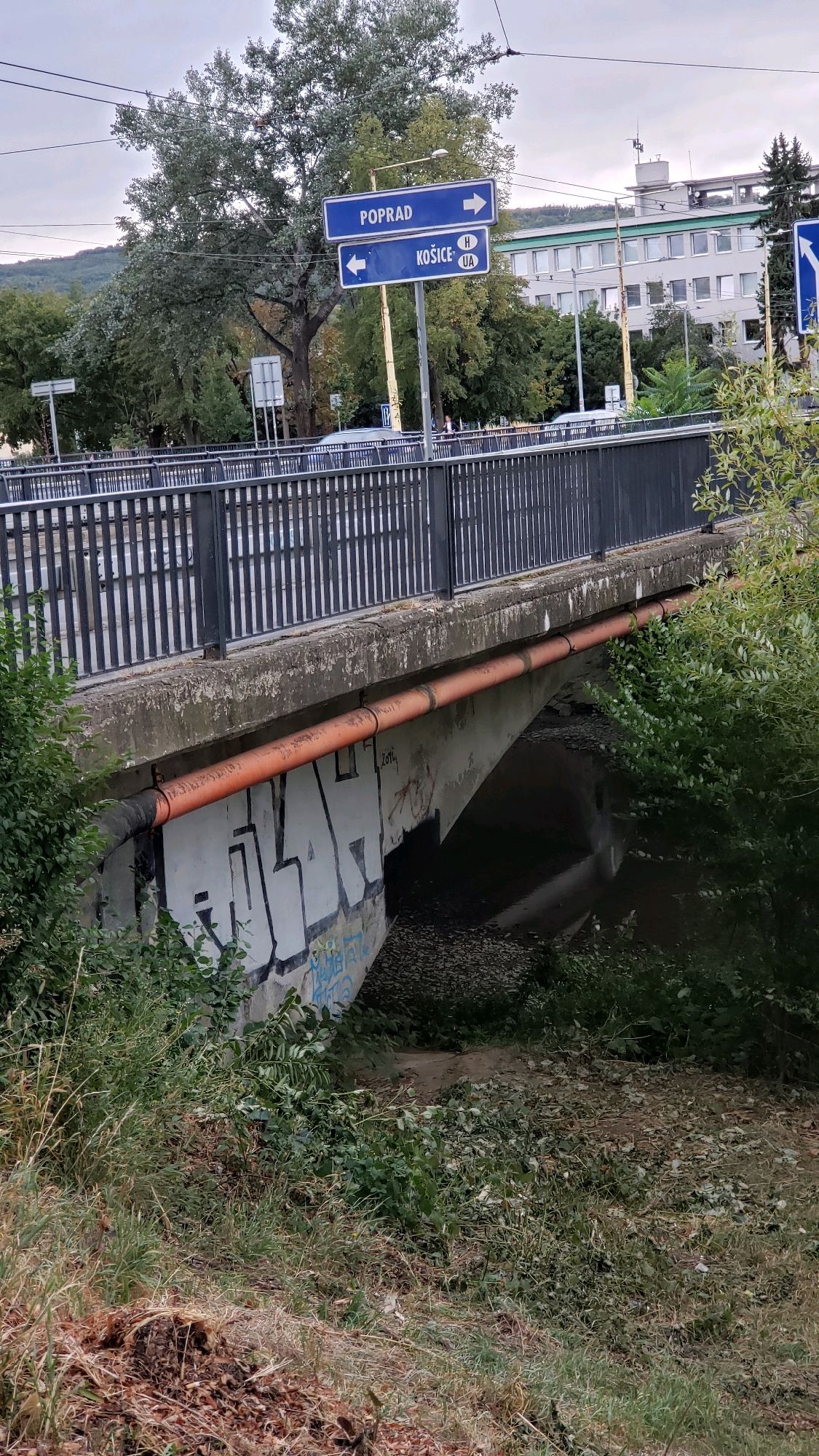 The image shows a concrete bridge with a metal railing over a dry streambed. There is graffiti on the underside of the bridge, and an orange pipe runs along the bottom edge. Two blue street signs are visible above the bridge, one pointing right with "POPRAD" and another pointing left with "KOŠICE" and flags of Hungary and Ukraine. The area around the streambed is overgrown with vegetation. A modern building is visible in the background, and trees line the street beyond the bridge. This location is on the road leading to the Nova Cvernovka industrial zone in Prešov, Slovakia, likely on the bridge crossing the L'ubovnianska street.