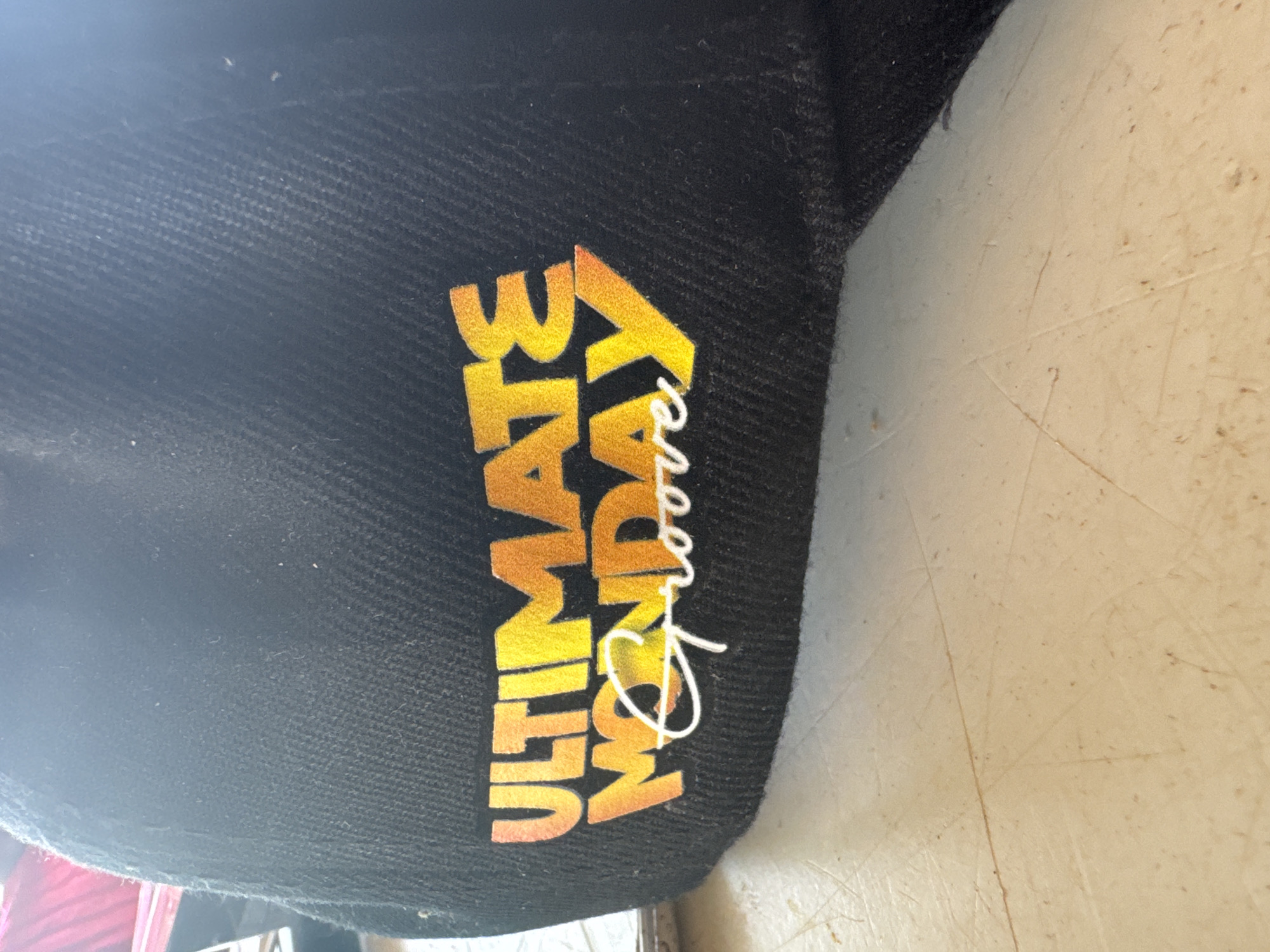 A black fabric item, likely a cap or hat, is prominently displayed in the image. The item features bold, multi-colored text on its surface. The phrase "ULTIMATE MONDAY" is rendered in a gradient block font, transitioning from orange at the top to yellow at the bottom. Overlapping the word "Monday" is the word "Groove" written in a stylized white script font. The fabric itself is a dark, possibly charcoal black, exhibiting a diagonal woven texture, and appears slightly dusty in some areas. The item rests on a light-colored, off-white surface that shows visible wear, including numerous scratches and small dark specks, suggesting it might be a table or workbench. The lighting is soft and even, without harsh shadows, indicating an indoor or shaded outdoor setting in Kurunduma, Nigeria. No people or specific activities are depicted; the image is a close-up still life focused on the apparel and its text.