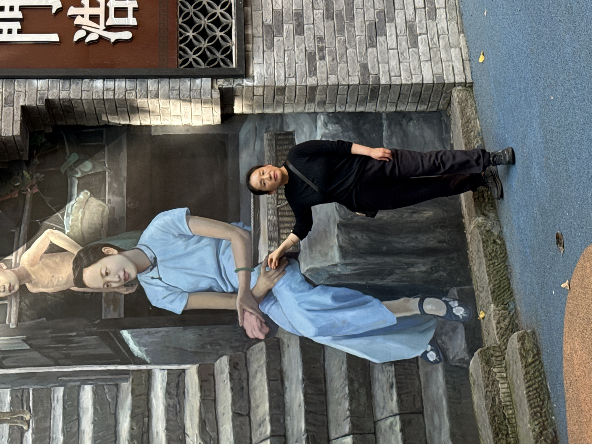 The image, oriented vertically, captures a woman posing alongside a detailed 3D street art painting. The woman, dressed entirely in black with a long-sleeved shirt, pants, and shoes, and carrying a black shoulder bag, stands on a blue textured ground. She is facing the viewer with a neutral expression, her right hand extended towards the painted scene, appearing to interact with it.
The street art itself depicts a realistic, deep illusion of a traditional or historical street. It features two prominent painted figures. One is a young woman in a light blue dress or tunic, with a green bracelet on her left wrist. Her black shoes are adorned with white floral embroidery. She is depicted looking downwards, her hands seemingly reaching towards a second painted figure who is partially visible, appearing emaciated and possibly lying or sitting. The painted background includes aged stone steps and dark buildings, evoking an old-world setting.
Above the painted art, real architectural elements are visible: a section of a gray brick wall, a brown panel with raised white Chinese characters that spell "琵琶巷" (Pípá Xiàng, meaning "Pipa Alley"), and a decorative black window grate with a geometric pattern. The scene is illuminated by natural daylight, creating shadows. Small yellow leaves are scattered on the blue ground where the real woman stands, adding a touch of natural detail to the outdoor setting. The image captures an engaging interaction between a person and immersive public artwork in Nan'an District, China.