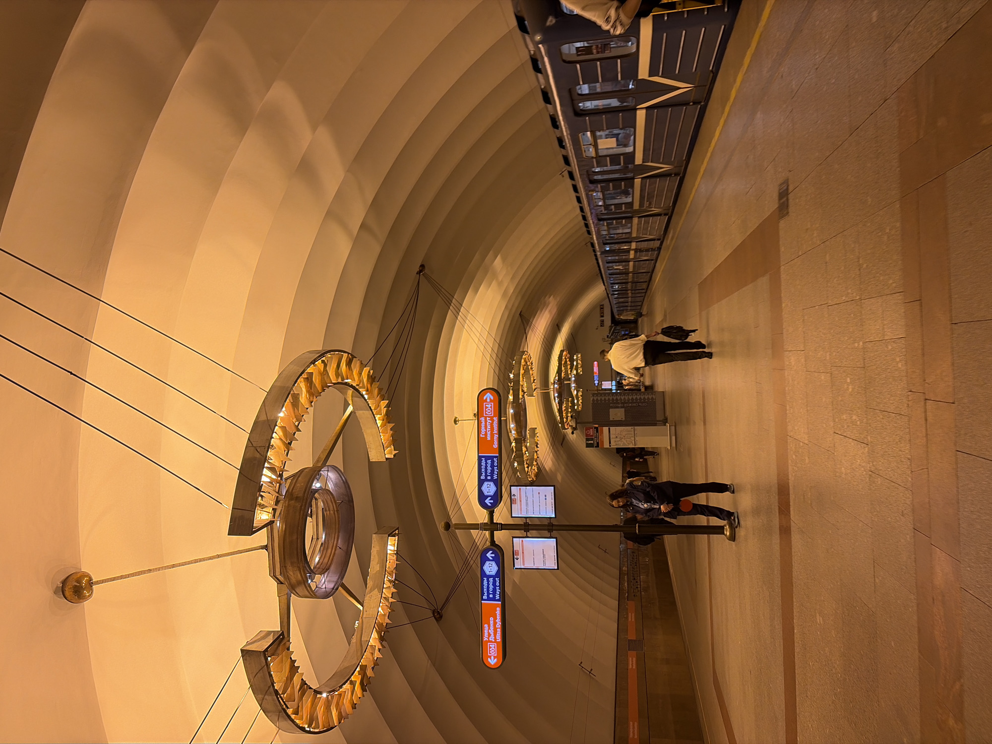 This is a photograph taken in a modern metro station, likely in Saint Petersburg, Russia, given the Cyrillic text. The scene is illuminated by warm, artificial lighting that casts long shadows, suggesting it's nighttime or underground. The station features a distinctive architectural design with a vaulted ceiling composed of repeating, curved panels that create a sense of depth and movement.
In the foreground, a large, abstract metallic sculpture with illuminated elements hangs from the ceiling. The platform itself is paved with large tiles and has a distinct line of orange tiles marking the edge. A signpost with directional arrows displays information in both Russian and English. The visible text indicates destinations such as "Ulitsa Dybenko" and "Gorny Institut," along with "Ways out" and platform numbers.
Several people are visible on the platform. One individual, dressed in dark clothing, is walking away from the camera, while another, wearing a light-colored jacket and dark pants, is also walking. Further down the platform, another person in dark attire is seen standing. The overall atmosphere appears calm and functional, characteristic of a public transportation hub. The image is captured with a wide-angle lens, contributing to the expansive and somewhat distorted perspective of the station's architecture.