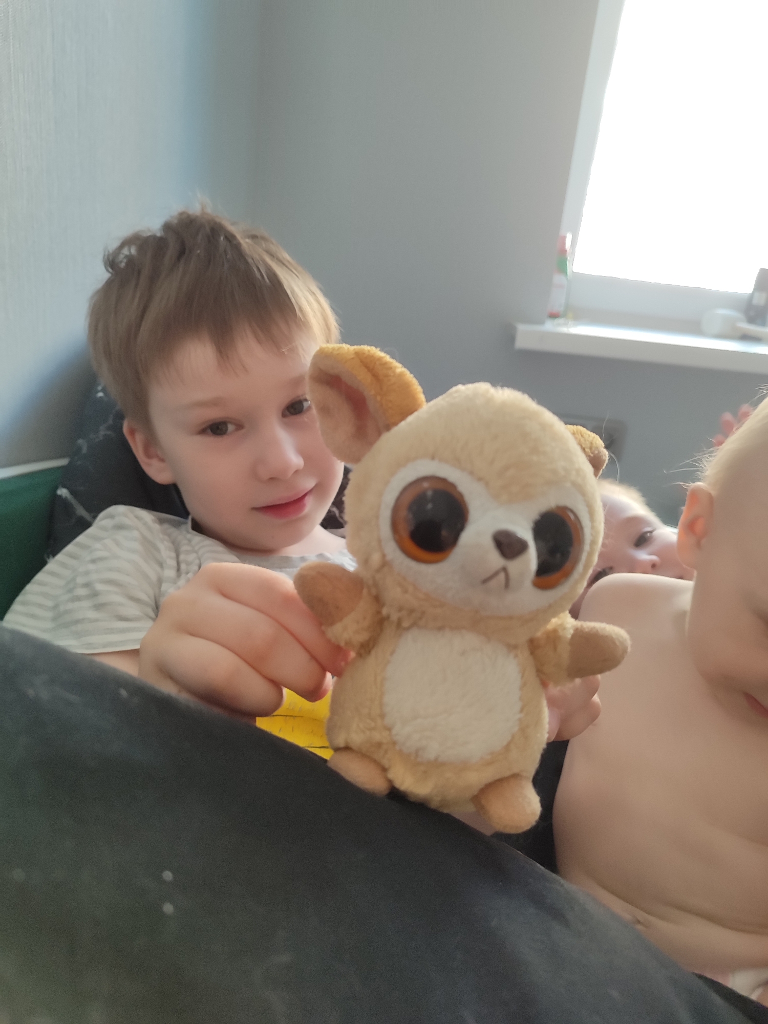 A medium shot shows a young boy holding a stuffed animal. The boy is fair-skinned with light brown hair and is wearing a striped gray and white shirt. He is smiling slightly and looking at the stuffed animal. The stuffed animal is a tan color with big brown eyes and is positioned in the center of the frame. In the background, partially obscured, are two other children. One is a toddler with fair skin and blonde hair, whose face is partially visible. Another child is seen from the side, revealing their bare torso and fair skin. The background includes a window with bright light shining through and a wall. The location is suggested to be Izhevsk, Russia, but there are no identifiable elements in the image to support or deny this claim definitively. The scene appears to be indoors, possibly a home setting.