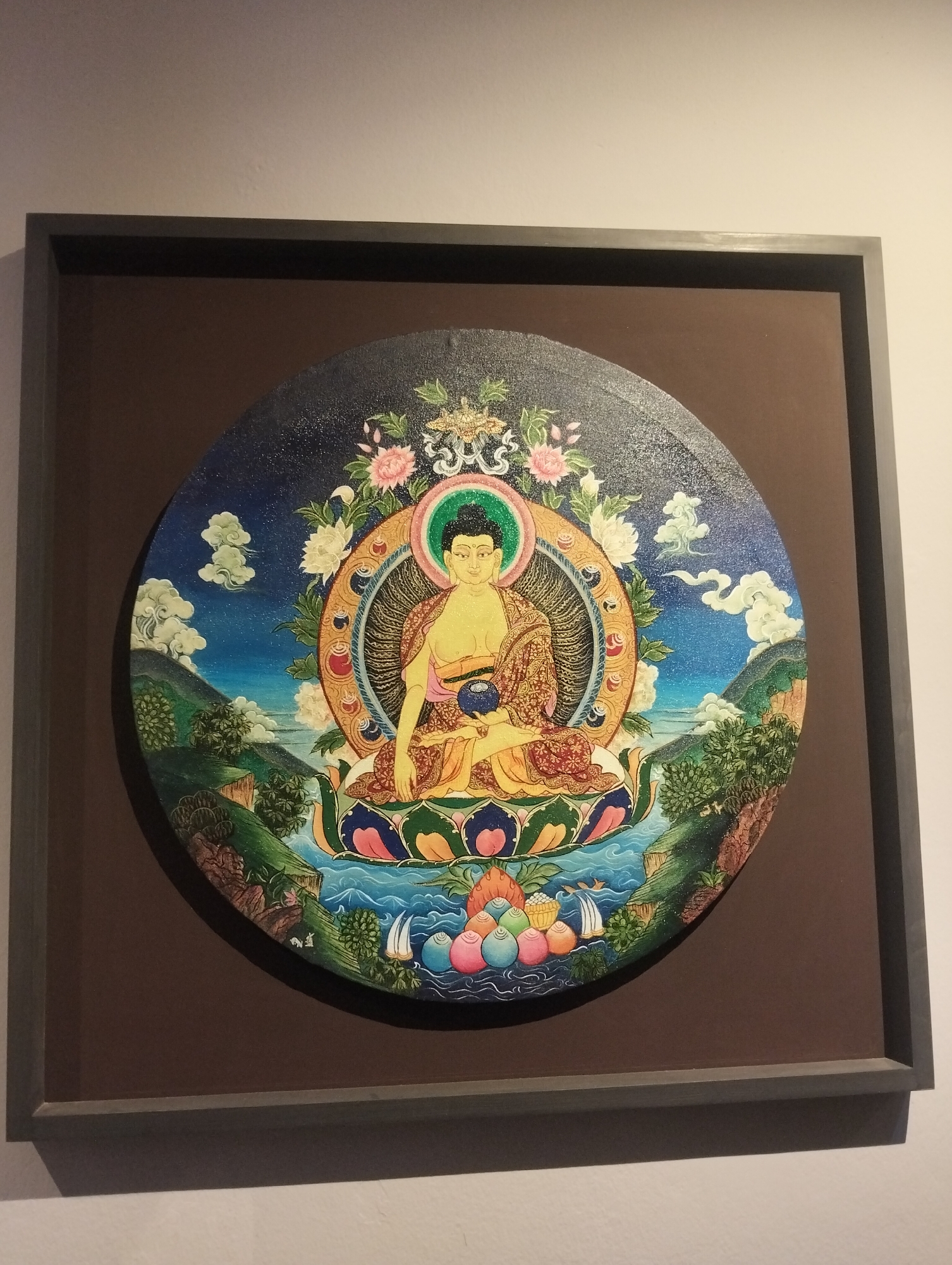 The image depicts a framed, circular painting of a Buddha-like figure seated on a lotus throne amidst a landscape. The figure has a green halo and is surrounded by flowers and a decorative aureole. The background shows mountains, clouds, and water. The painting is mounted on a brown background within a dark grey frame. The entire piece is displayed on a light-colored wall. The city and location of the image were not stated.