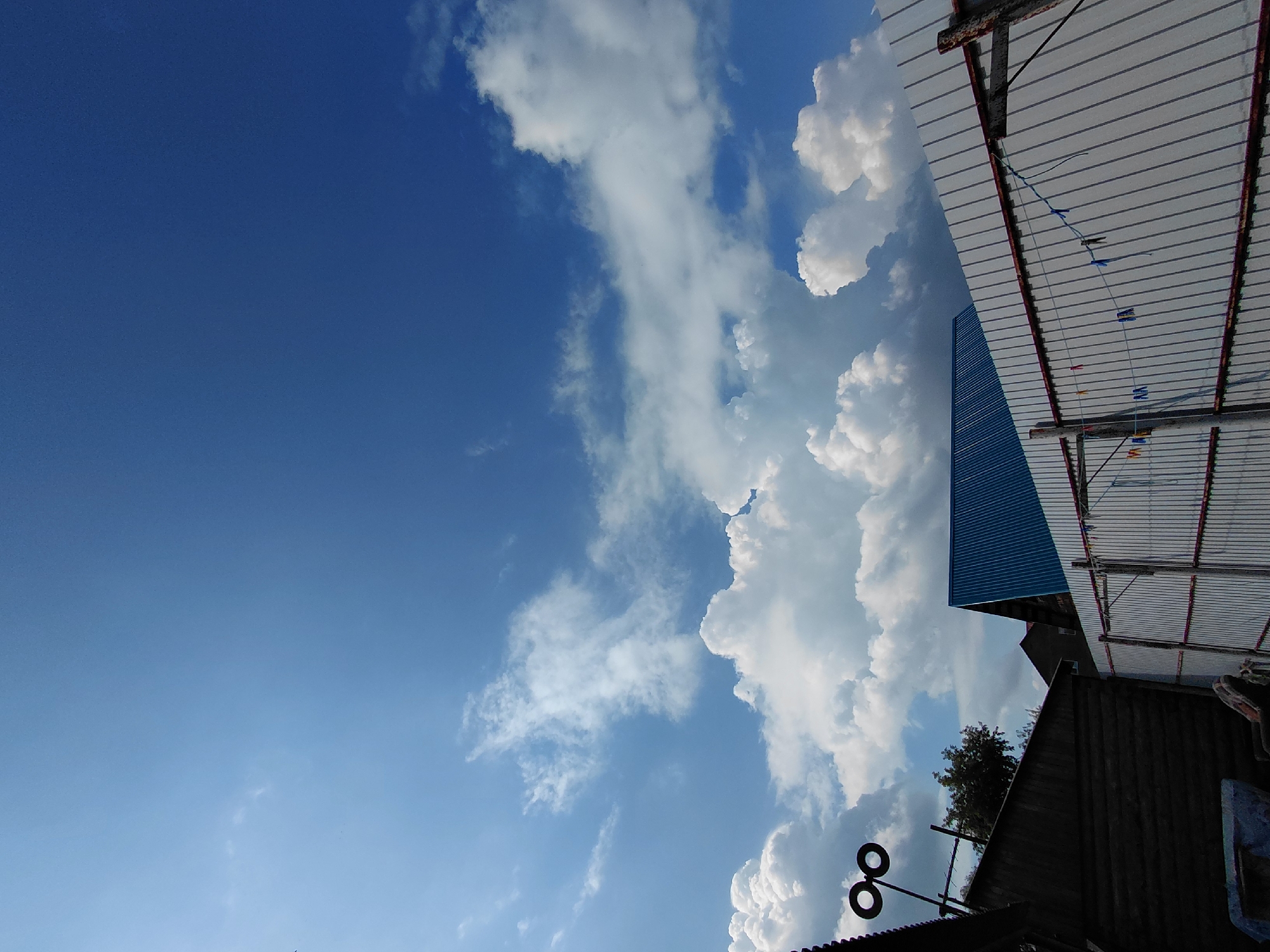 The image depicts a clear blue sky with scattered white clouds, characteristic of a summer day. In the foreground, the rooftops of several buildings are visible, including one with a blue corrugated metal roof and another constructed of dark wooden logs. A white corrugated metal fence runs across the bottom right of the image, with a clothesline strung between two posts, adorned with several colorful clothespins and some hanging laundry. To the left, a dark structure with a slanted roof is visible, and above it, two black circular objects are mounted on a pole. The overall scene suggests a residential or semi-rural area. This image was taken in Novosibirsk, Russia.