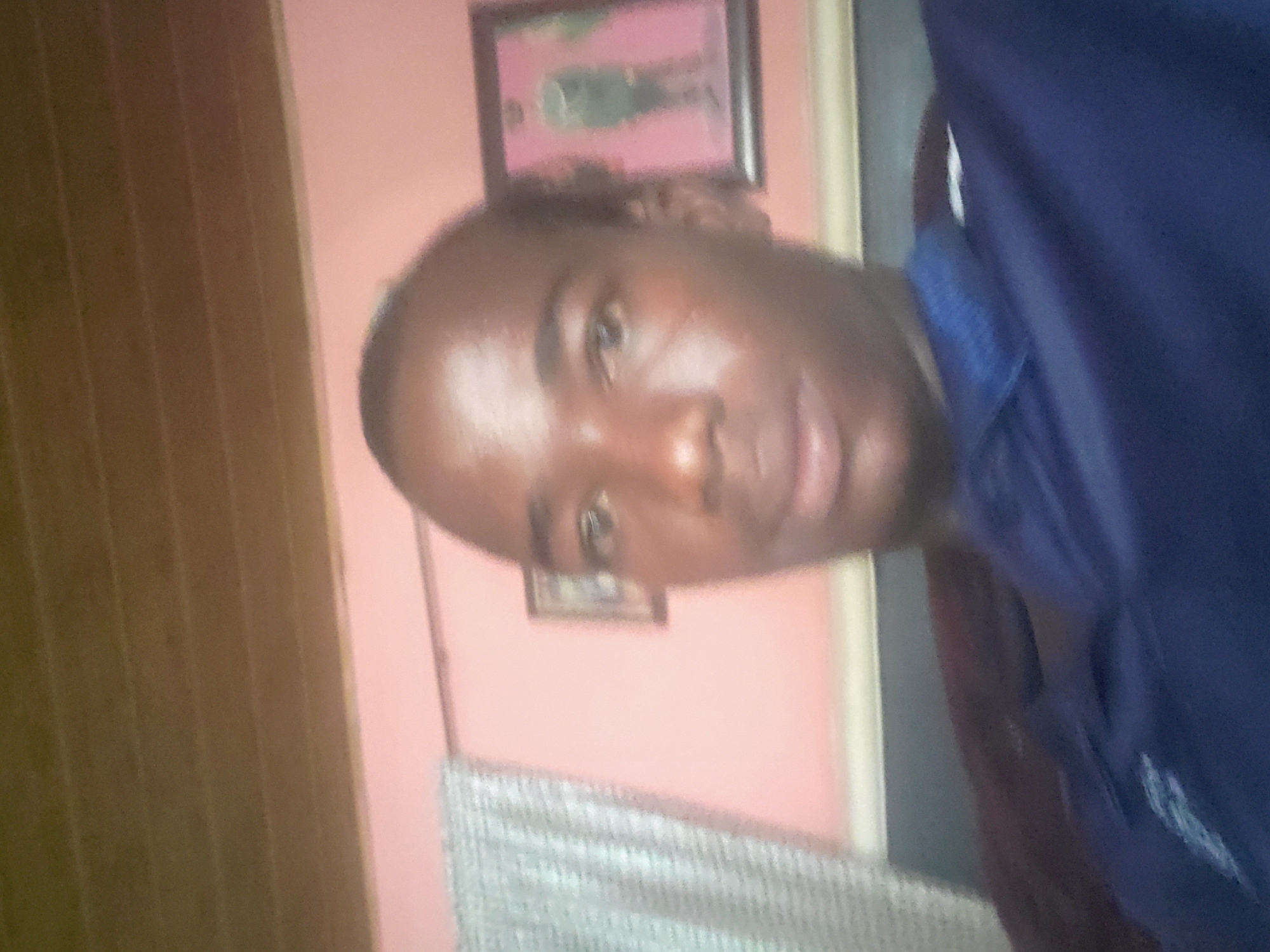 The image is a close-up, blurry selfie of a young Black male wearing a navy blue polo shirt. He is positioned in the center of the frame, with his face slightly turned to the right. The background consists of a pale pink wall, a framed picture on the wall depicting a person in uniform, and a section of a wooden slatted ceiling. To the left of the frame, a light-colored, textured curtain is visible. The lighting appears to be from an overhead source, casting highlights on the subject's face. The overall impression is of an indoor setting, likely a residential or personal space. Without further context or visible landmarks, it is not possible to pinpoint the exact location within Oturkpo, Nigeria.