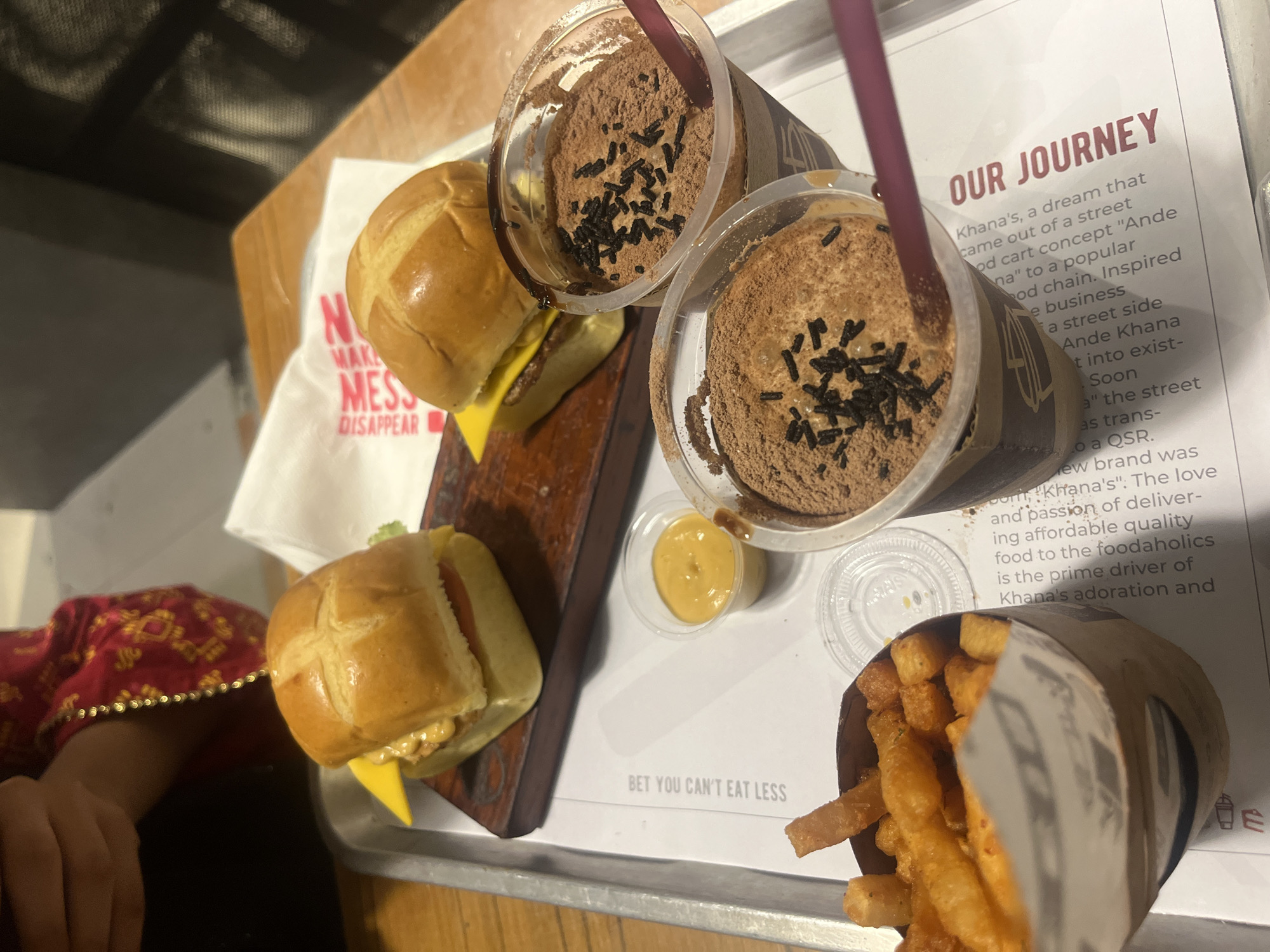 The image shows a meal on a tray, possibly at a restaurant in Dhaka, Bangladesh. The tray contains two small burgers served on a wooden plank, two chocolate milkshakes with sprinkles, a container of sauce, and a cone of french fries. A white paper on the tray with the words "BET YOU CAN'T EAT LESS" and "OUR JOURNEY" printed is also visible, along with a brief description of "Khana's". A person's arm and hand in a red and gold garment is visible in the lower left corner.