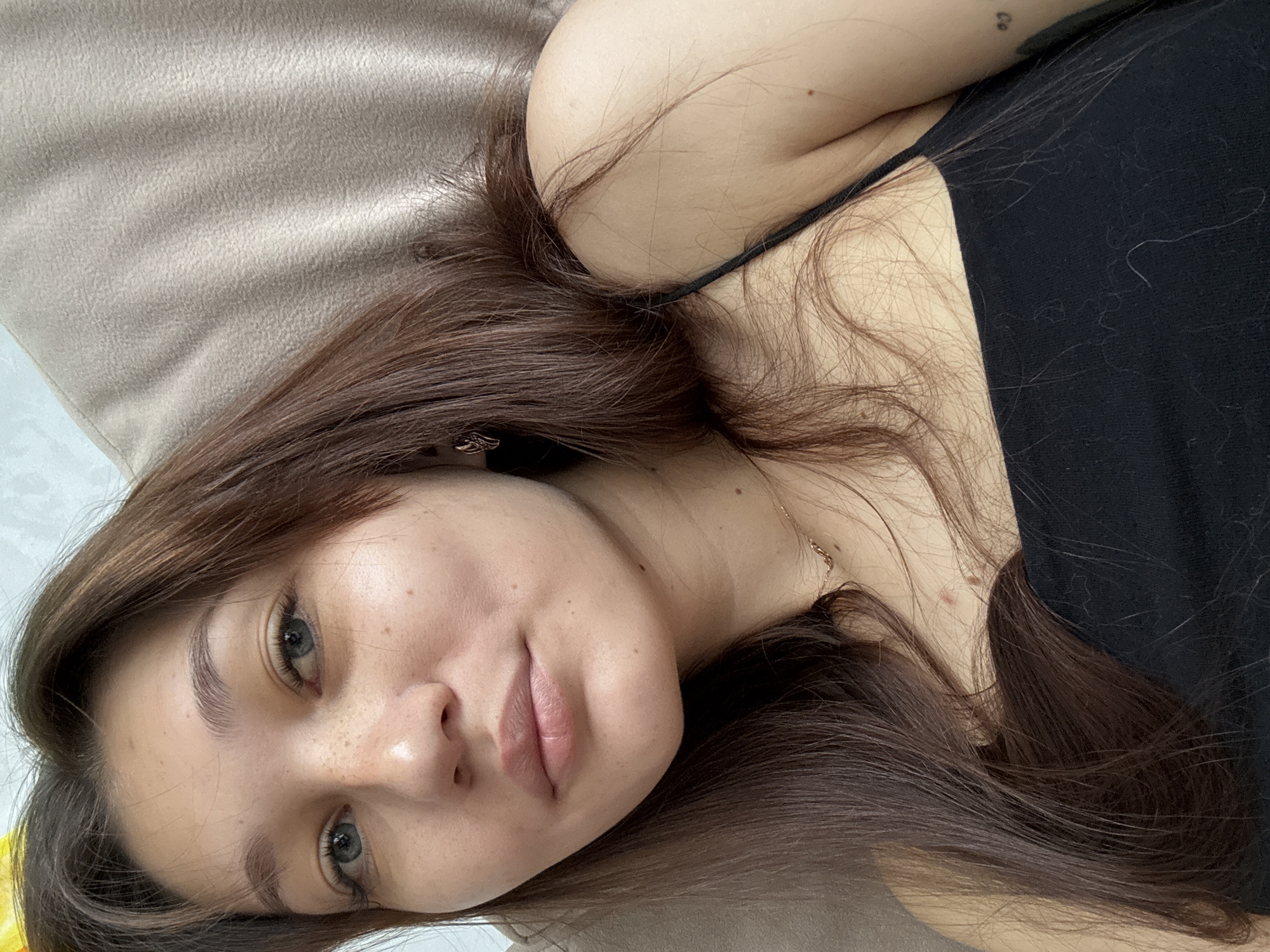 The image is a close-up, vertically oriented, selfie-style shot of a young woman with long, dark brown hair. She is lying down, with her head resting on a textured, light-brown surface, possibly a pillow or blanket. Her gaze is directed towards the camera, and her expression appears relaxed or pensive. She is wearing a black tank top, and a thin gold chain is visible around her neck. Her skin is fair with some freckles visible on her cheeks and nose. The lighting suggests it is daytime, possibly indoors given the soft light and textures. The background is out of focus, but a light-colored surface is visible to the left. There are no visible text elements in the image. The overall mood is intimate and casual.