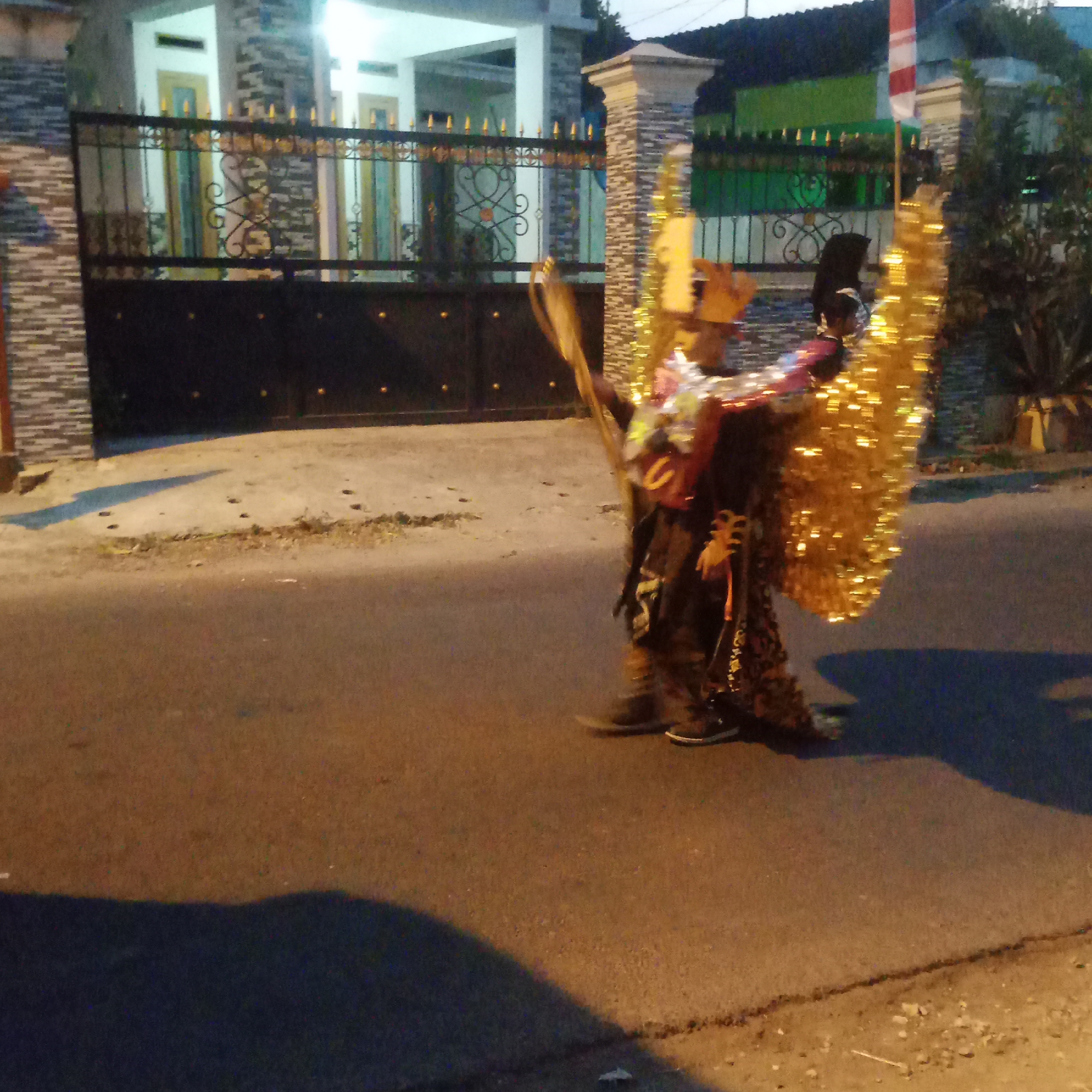 The image depicts a street scene at dusk or night in Semanding, Indonesia. Two individuals are visible, one in the foreground on the right and another partially obscured behind them. The person in the foreground is wearing an elaborate costume that includes large, shimmering golden wings. Their attire appears to be a combination of fabric, possibly with reflective elements, and ornate detailing. They are walking on a paved street. Behind them, a second individual, dressed in a black hijab and possibly a dark-colored garment, is visible, facing away from the camera. They seem to be interacting with something in their hands, though the details are unclear.
The background reveals a residential area. A dark metal gate with decorative gold accents is prominent, behind which a house with white pillars and patterned walls can be seen. The house appears to have multiple entrances. To the right of the house, a section of a green building with a white and red Indonesian flag is visible. The street in front of the house is paved with asphalt and shows some gravel and debris along the edge. Shadows are cast on the street, indicating artificial lighting or the fading daylight. The overall atmosphere suggests a festive or ceremonial event taking place in the neighborhood.