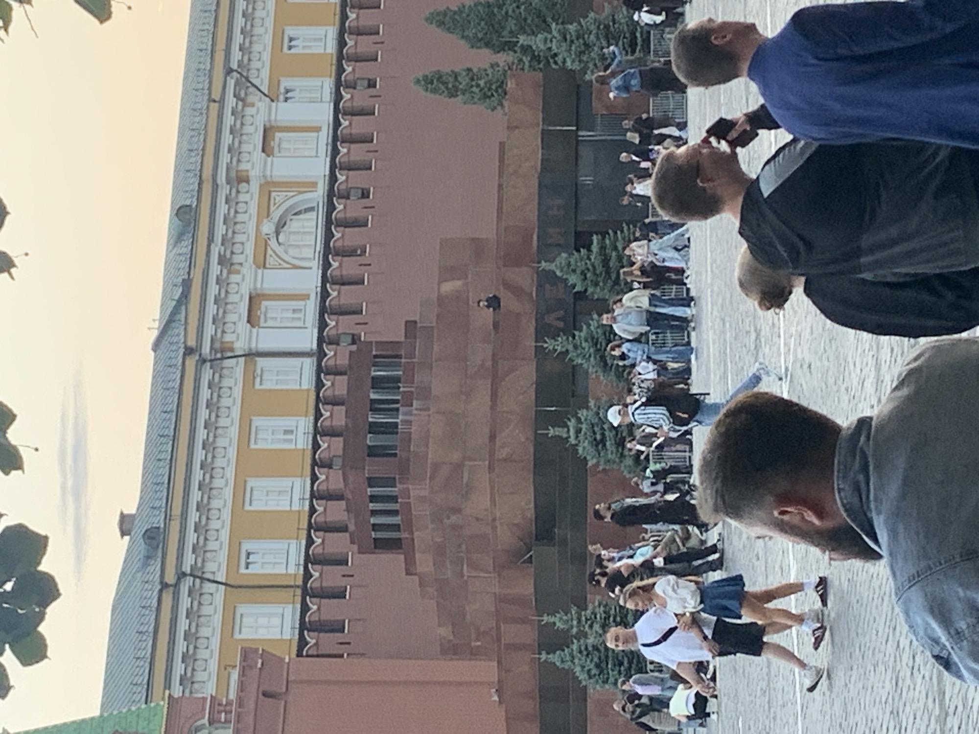 The image depicts Red Square in Moscow, Russia. In the foreground, several people are visible from behind, some walking and others standing. A man in a grey denim jacket is in the closest foreground, looking to his right. To his left, a man in a black hoodie is holding a dark object to his ear, possibly a phone. To the right of the man in the hoodie, a man in a blue sweater is facing away from the camera.
In the midground, a diverse group of people are walking and standing. A couple, a man in a white t-shirt and black shorts and a woman in a white top and blue skirt, are walking towards the left. Behind them, a line of people are seen walking and standing near a fence. Pine trees are scattered throughout this area.
The background is dominated by the Lenin Mausoleum, a stepped, tiered structure made of red brick. Behind the mausoleum, a yellow building with white decorative elements and windows can be seen. The sky above is a pale orange, suggesting it is either sunrise or sunset. The ground is paved with cobblestones.