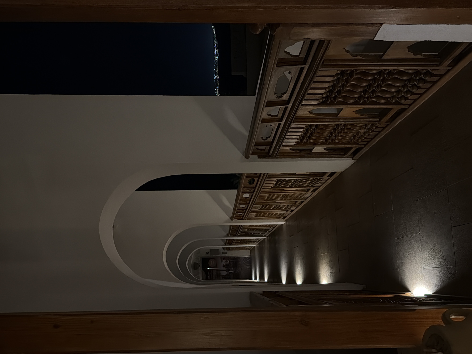 The image depicts a tunnel-like space in Sharm El Sheikh, Egypt. White walls with rounded archways line one side, illuminated by recessed lights. The opposite side features dark tiled walls also with recessed lights. A decorated wooden structure runs along the top of the tiled wall. Outside the tunnel, a glimpse of the city at night is visible. Wooden beams appear at the top and bottom of the frame.