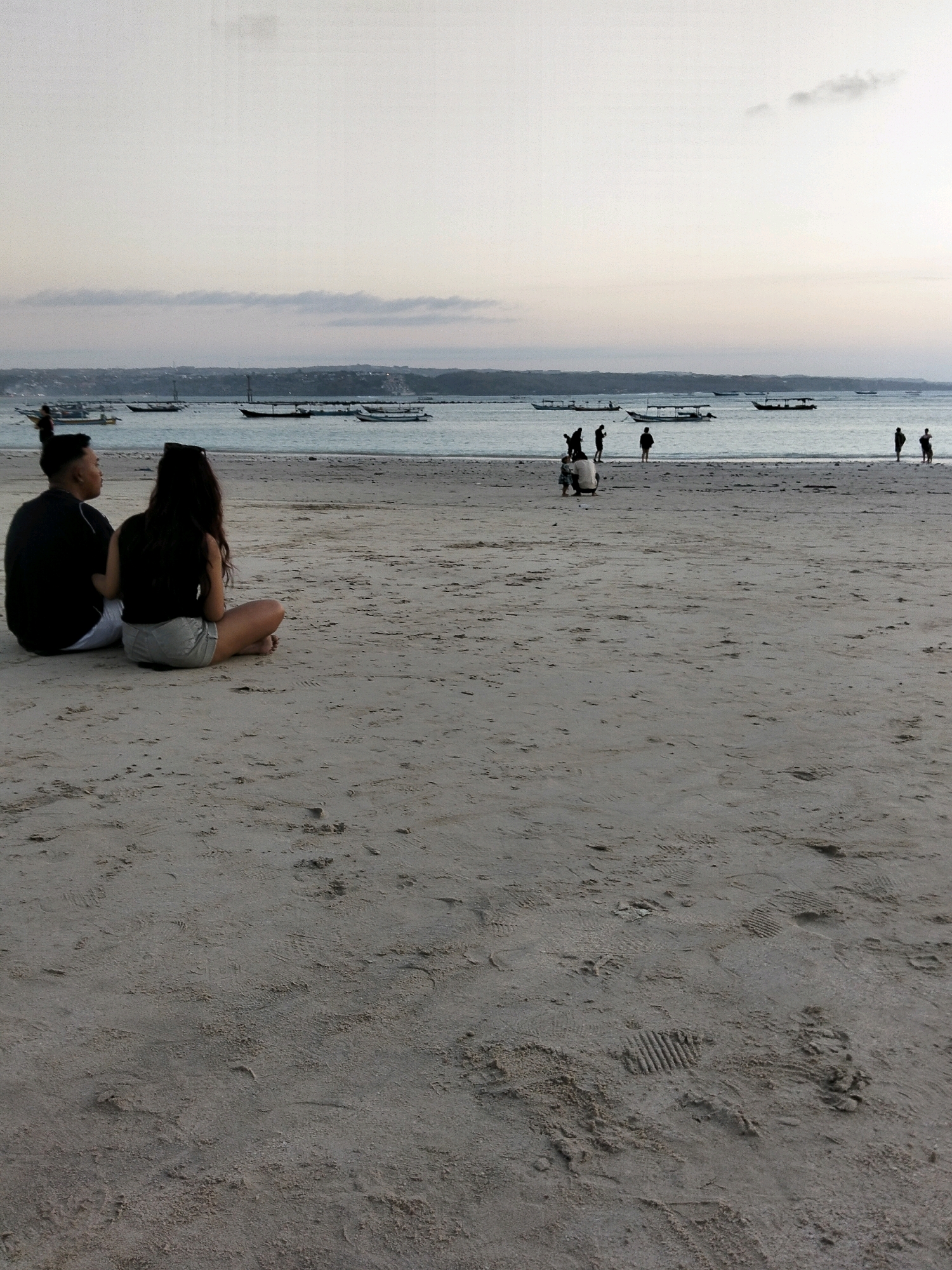 In Kuta, Indonesia, a serene beach scene unfolds at dusk. Two individuals, a male with hair styled in a bun and a female with long dark hair, sit on the sandy shore, facing the ocean. They are dressed casually, with the male in a black t-shirt and the female in a black top and grey shorts. Their posture suggests a relaxed moment of observation.
Further along the shoreline, several other figures are scattered, some standing and others appearing to be engaged in activities near the water's edge. In the distance, a line of boats rests on the calm sea, silhouetted against the pale, hazy sky. The horizon is marked by a faint landmass, possibly a coastline or distant hills. The sand itself is textured with footprints and subtle undulations, indicative of a well-trodden public beach. The overall atmosphere is one of quiet tranquility as the day transitions into evening.