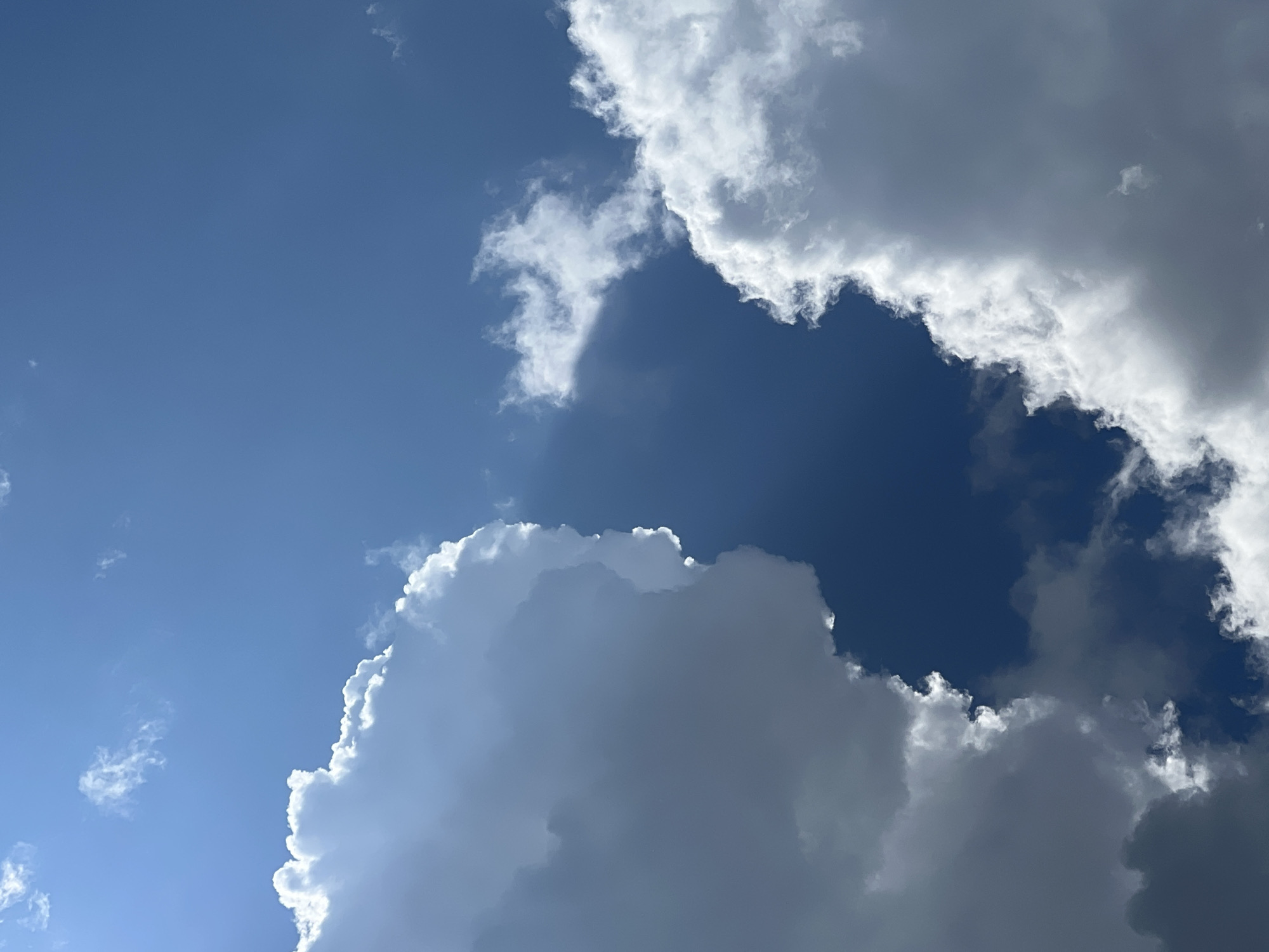 The image depicts a segment of the sky with prominent cloud formations and a clear blue expanse. The clouds are varied, with some appearing bright white and fluffy, catching the sunlight, while others are darker, suggesting depth or thickness. A shaft of sunlight appears to be breaking through one of the clouds, creating a visible ray. The scene is devoid of any human presence, structures, or terrestrial elements, focusing solely on the atmospheric conditions. The sky is a deep blue, indicative of a clear day.