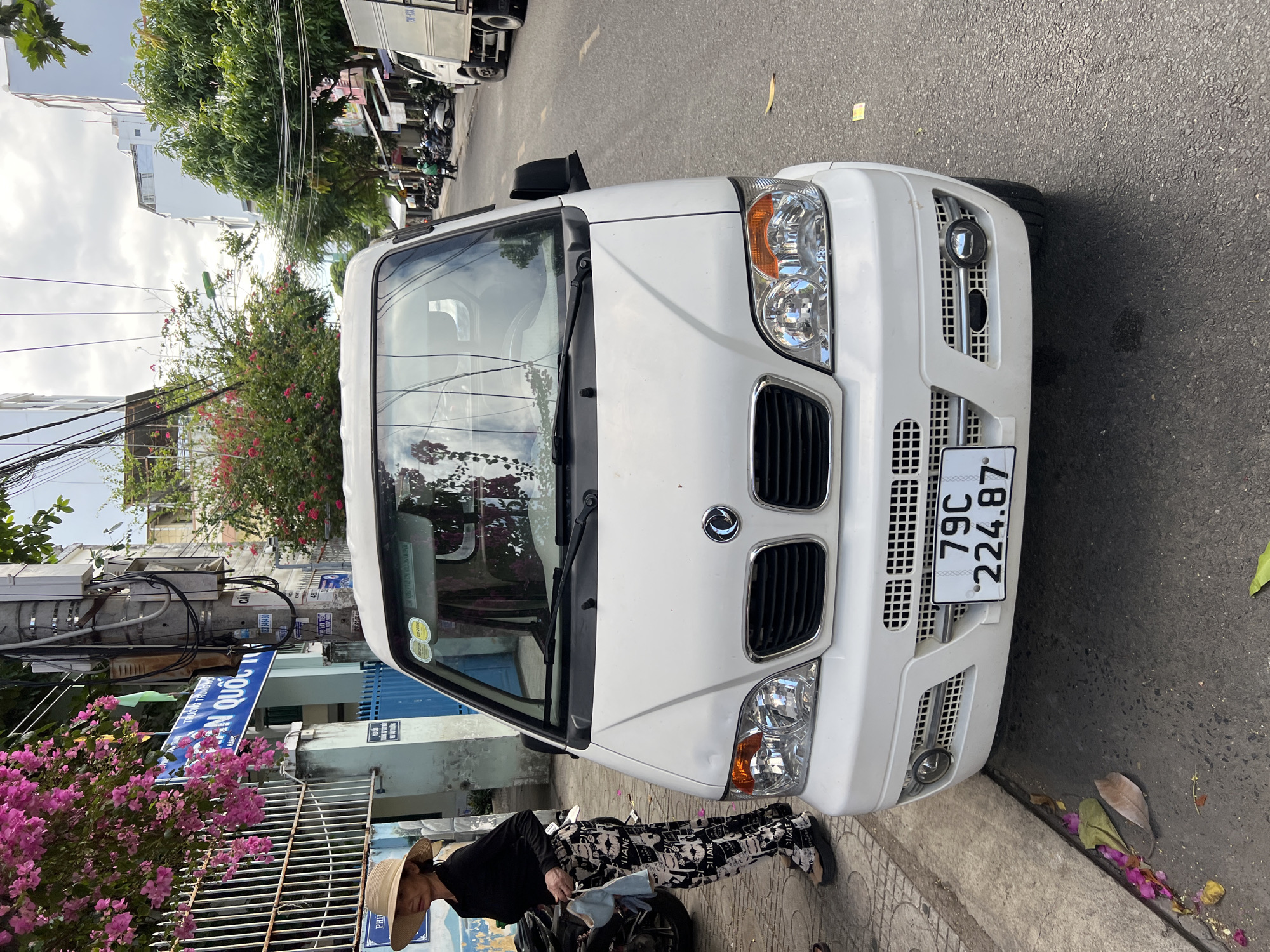 A white van is parked on a street in Nha Trang, Vietnam, facing the camera. The van has a license plate reading "79C 224.87". A woman wearing a wide-brimmed hat, a black long-sleeved shirt, and patterned pants stands on the sidewalk to the left of the van, holding a blue cloth. Pink bougainvillea flowers hang from a nearby fence. The street is paved, and there are buildings, power lines, and other vehicles visible in the background. The sky is overcast.