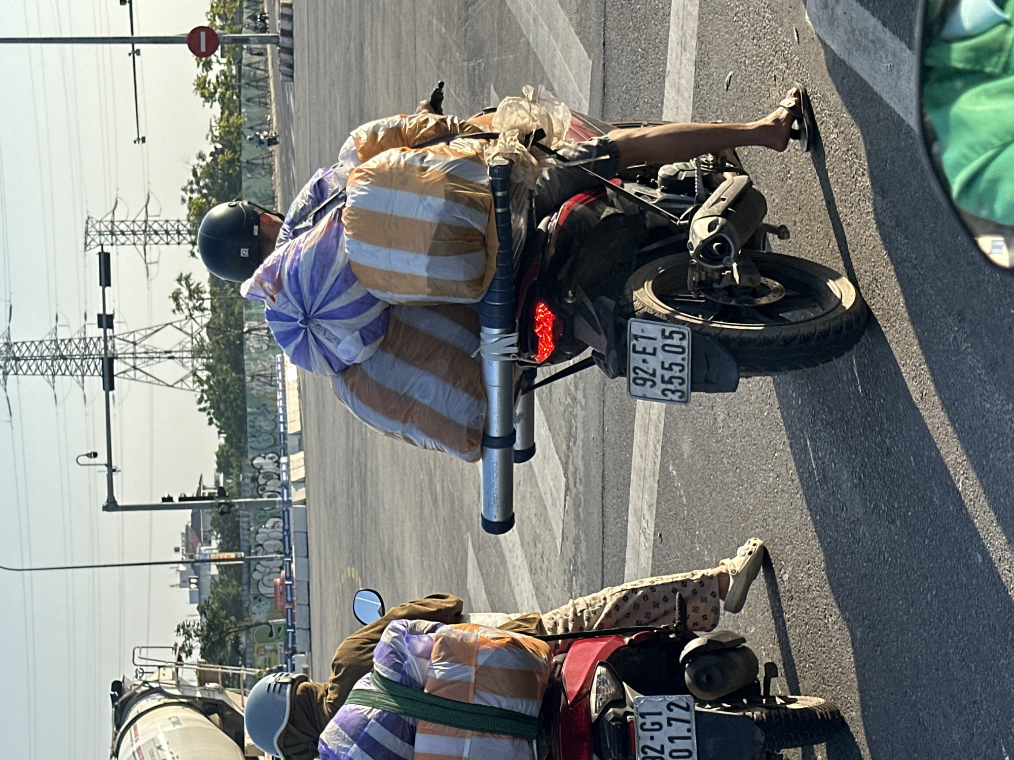 Two motorbikes are stopped side-by-side on a paved road in Đà Nẵng, Vietnam. The first motorbike is burgundy, and the second is black. Each bike is heavily laden with striped bags, secured on the back. The riders both wear helmets, with the first rider wearing light tan pants, and the second rider wearing dark shorts. License plates are visible on both bikes, with the first reading "92-G1 01.72" and the second reading "92-E1 355.05". In the background, power lines and buildings are visible. There is a cement truck near the first bike and a street sign above the second bike.