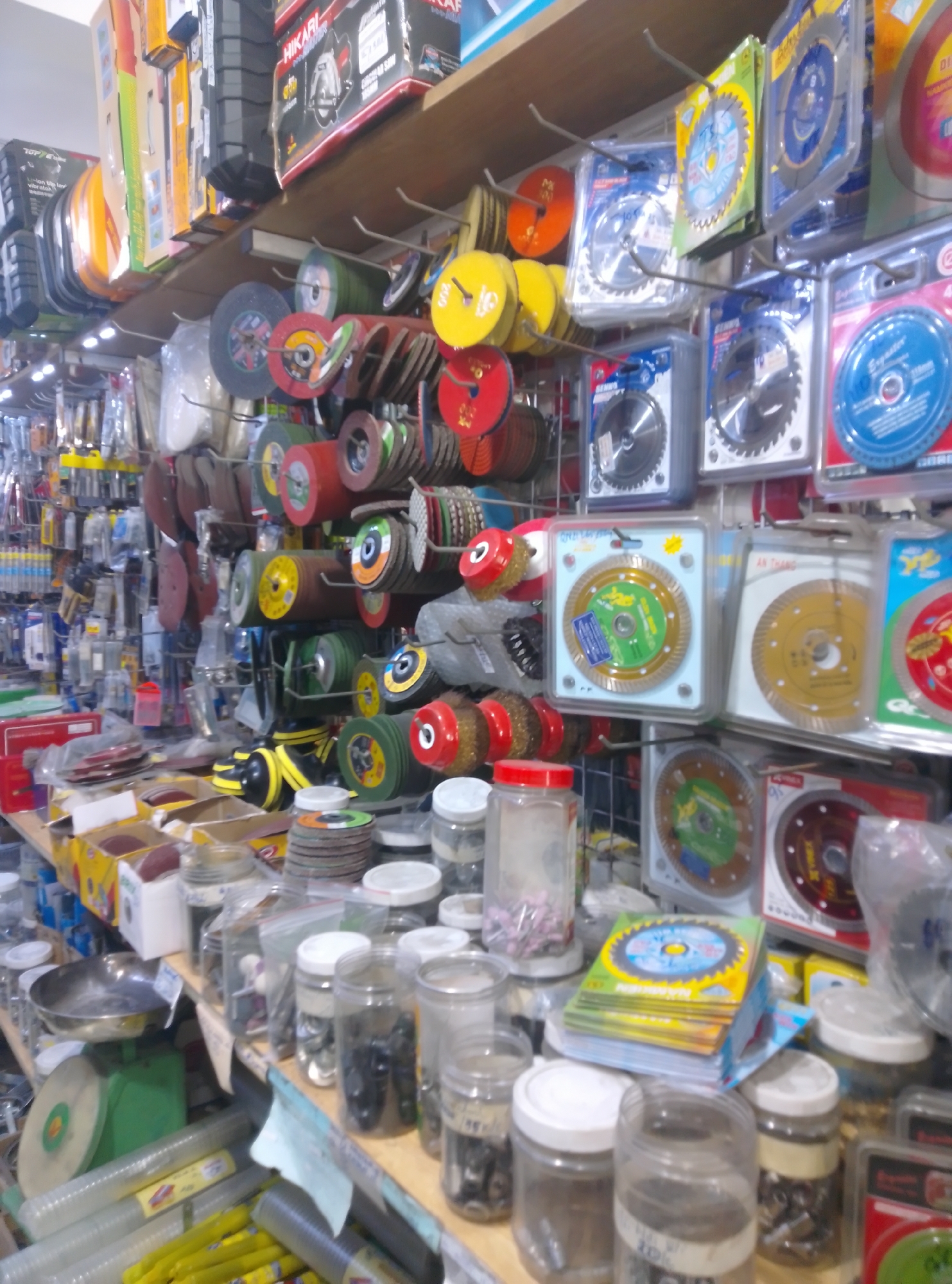 This image captures the interior of a densely stocked hardware or tool supply store located in Bac Nha Trang Ward, Vietnam. No individuals are present in the scene. The store features a functional layout with wooden shelves along the top and extensive metal grid display racks extending vertically, showcasing a vast array of tools and accessories.
The dominant subjects are abrasive tools and cutting blades. Numerous grinding wheels, flap discs, and cut-off wheels are displayed in various colors (red, green, yellow, brown, black) and sizes, often stacked or hanging. Prominently featured are circular saw blades for different applications, including toothed blades and diamond blades, packaged in clear plastic and suspended from hooks. Partially visible brand names include "SENHS," "HIKARI," and "AN THANG."
Lower sections of the store contain additional merchandise: clear plastic jars hold small hardware components, such as bolts and grinding points (some pink-colored); bundles of yellow-wrapped sandpaper and clear plastic tubing are visible; and cardboard boxes filled with what appear to be sanding blocks or similar items. A green analog weighing scale with a metal pan is also discernible on a lower surface. The environment is that of a bustling retail outlet, heavily focused on industrial or construction supplies, illuminated by overhead artificial lighting.