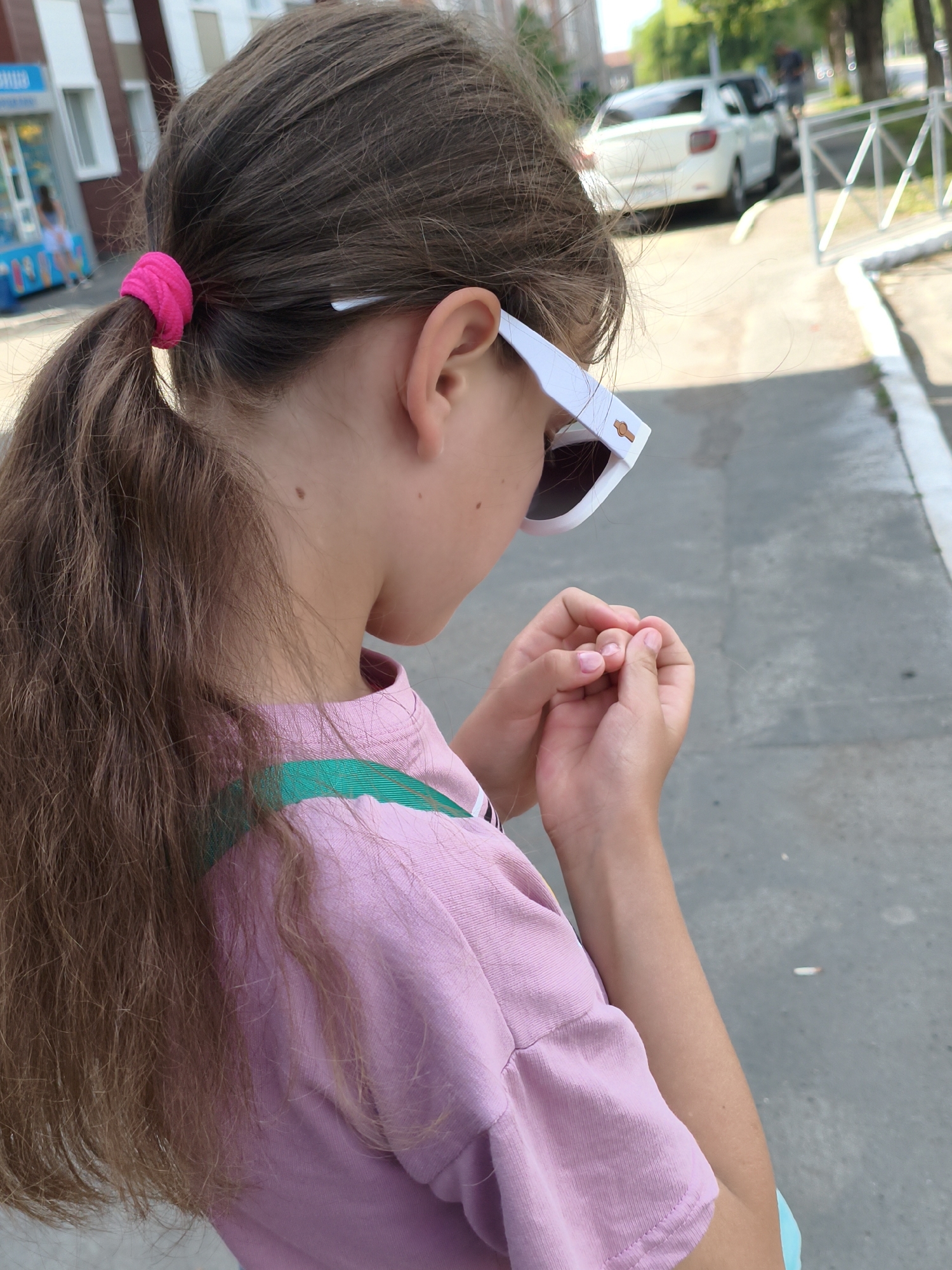 The image shows a young girl from the side, wearing a pink t-shirt and white sunglasses with a stylized watch design on the frames. Her long, brown hair is pulled back into a ponytail with a bright pink scrunchie. She is holding her hands clasped in front of her. The background suggests an outdoor urban environment, with a building to the left and a street with a white car and a metal fence to the right. The lighting indicates it is daytime, likely sunny. The image appears to be taken on a sidewalk. The girl's attention is focused downwards on her hands.