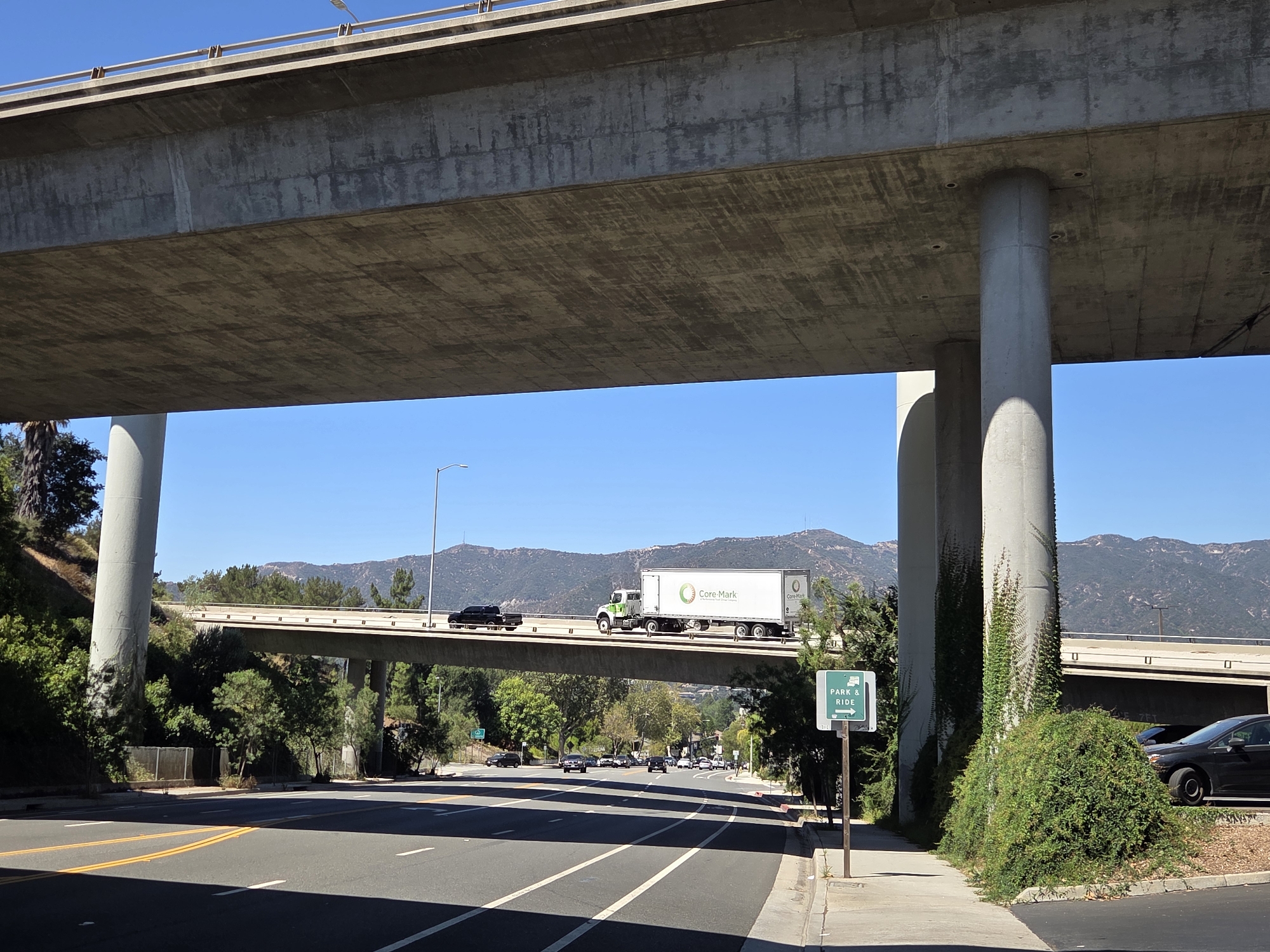 This is a full, eye-level, outdoor shot of an elevated highway overpass in Glendale, United States of America, on a clear day. The overpass is constructed from concrete and supported by large cylindrical columns. A white semi-trailer truck with "Core-Mark" branding is driving on the overpass. Several other vehicles are also visible on the overpass and in the distance on a lower road. Green trees and mountains form the background. A "PARK & RIDE" sign with an arrow pointing to the right is positioned on the right side of the foreground. The foreground shows a paved road with lane markings.