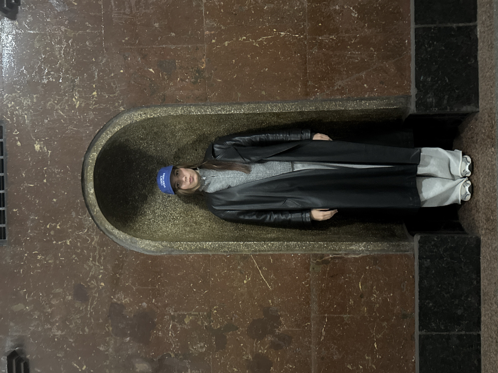 A woman wearing a blue hat, gray sweater, black coat, and gray pants is lying in a disused public drinking fountain. The fountain is set into a wall constructed of brown and black stone and is likely located in the Moscow subway.