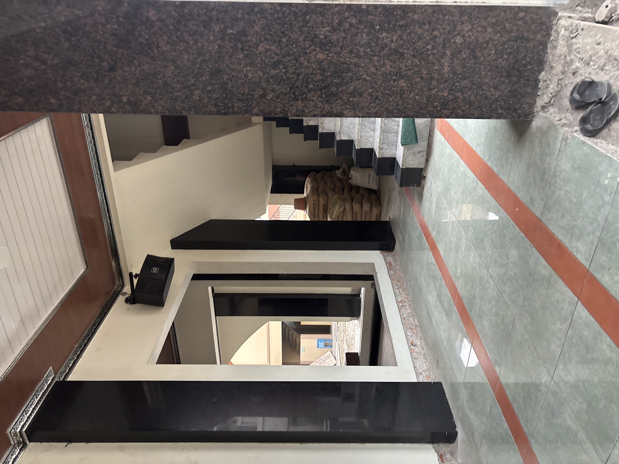 This image depicts an indoor architectural space, possibly a building lobby or entryway, located in Bekasi, Indonesia. The flooring is tiled with a light green and grey pattern, accented by two parallel orange stripes running horizontally across the image. To the left, a tall, dark, polished column stands prominently, contrasting with the lighter colored walls. Further left, a large dark framed opening reveals another area with an arched passageway leading to a staircase and what appears to be an outdoor or courtyard-like space.
In the foreground, a dark speaker is mounted on the wall above the opening. To the right of the image, another large, dark, granite-patterned column dominates the view. Behind this column, a set of marble stairs with dark risers ascends. Stacks of large, burlap sacks, likely containing building materials or commodities, are visible near the base of the stairs. Two dark flip-flops are resting on the ground in the lower right corner of the frame, near the base of the granite column. The ceiling is white and appears to be paneled, with a brown border trim along the edges of the main ceiling area. The overall impression is of an unfinished or utilitarian space with elements of modern construction and materials.