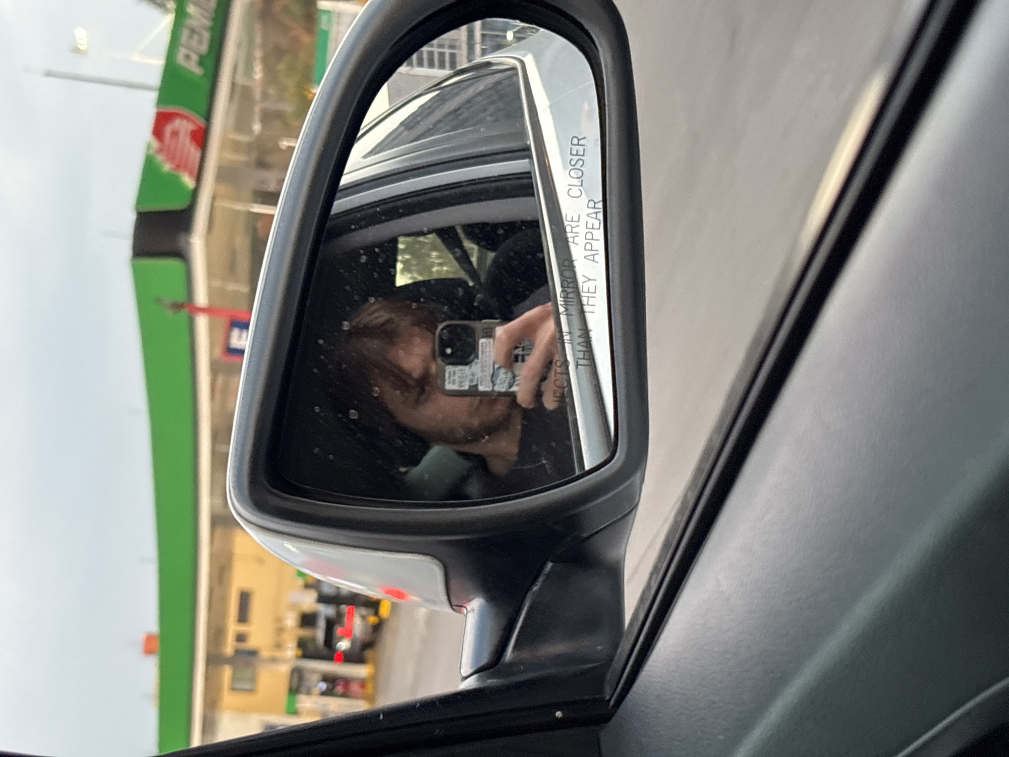 The image shows a car's side mirror, reflecting the driver taking a photo of themselves with a smartphone. Outside the car window, a Pemex gas station is visible. This location is in Huixquilucan de Degollado, Mexico.