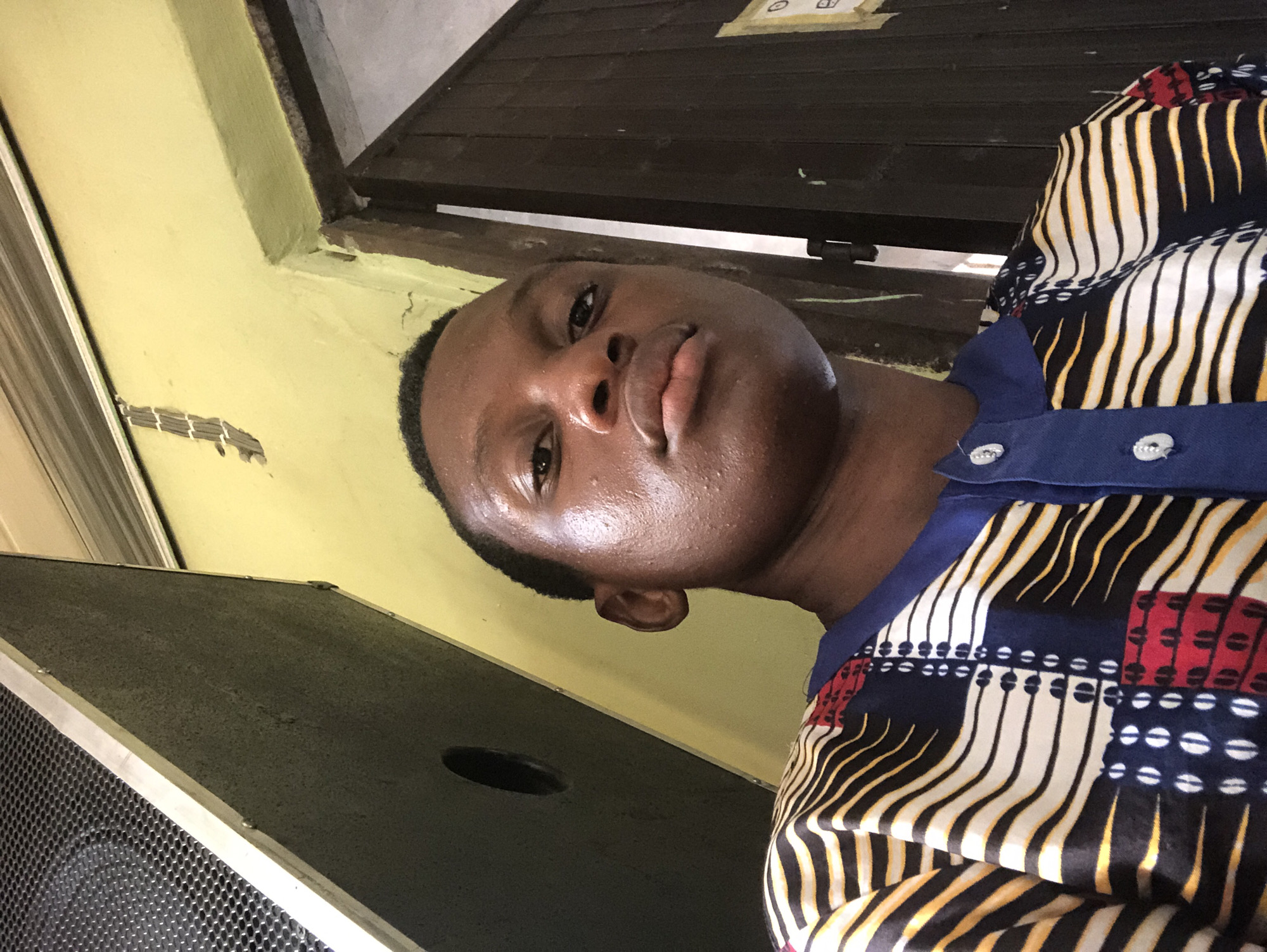 A medium-close selfie of a young Black male is oriented such that his head is at the top of the frame and rotated slightly to the left. His skin appears slightly damp or oily. He is wearing a button-down shirt with a dark blue collar and a pattern of vertical stripes in white, black, and yellow, interspersed with sections of red and blue featuring white oval shapes.
The background includes a light yellow wall with some cosmetic damage, a dark brown wooden structure above the wall, and the corner of a large black speaker to the left. The photo was taken indoors. Specific location in Ukwu-Abwa, Nigeria, cannot be determined.