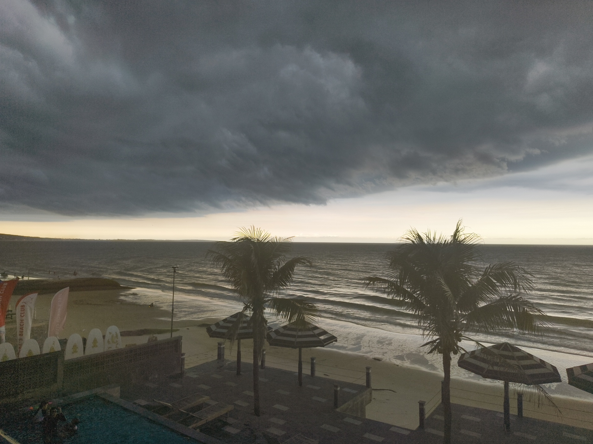 The image depicts a beach scene with a large, dark storm cloud dominating the sky over the ocean. The waves are visible on the sandy beach, and there are several palm trees and striped umbrellas on the shore. In the foreground, a swimming pool with people in it is partially visible. Flags and surfboards are placed along the beach area. The overall atmosphere is overcast and stormy, suggesting impending rain. The location is identified as Phu Thuy Ward, Vietnam.
