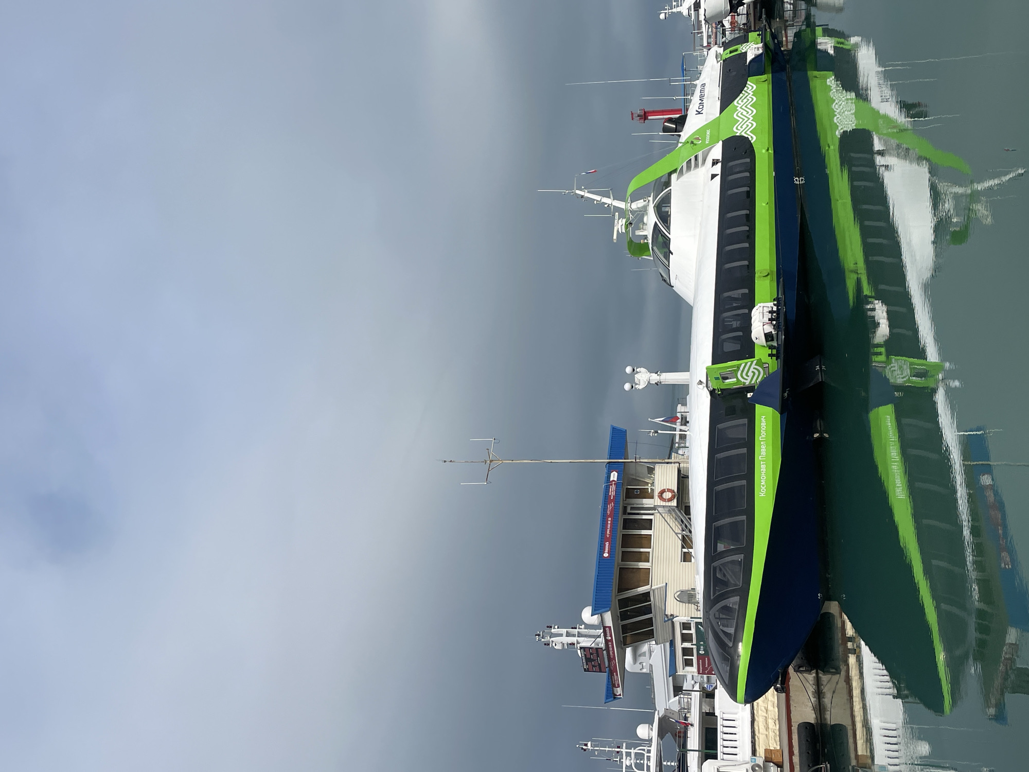 The image shows a hydrofoil vessel, "Kometa," docked in Sochi, Russia. The vessel is painted white, dark blue and green, and the words "Kometa" are visible on the vessel. The vessel is in calm water, which reflects the vessel's image. Several other boats are docked nearby. The sky is overcast and grey.