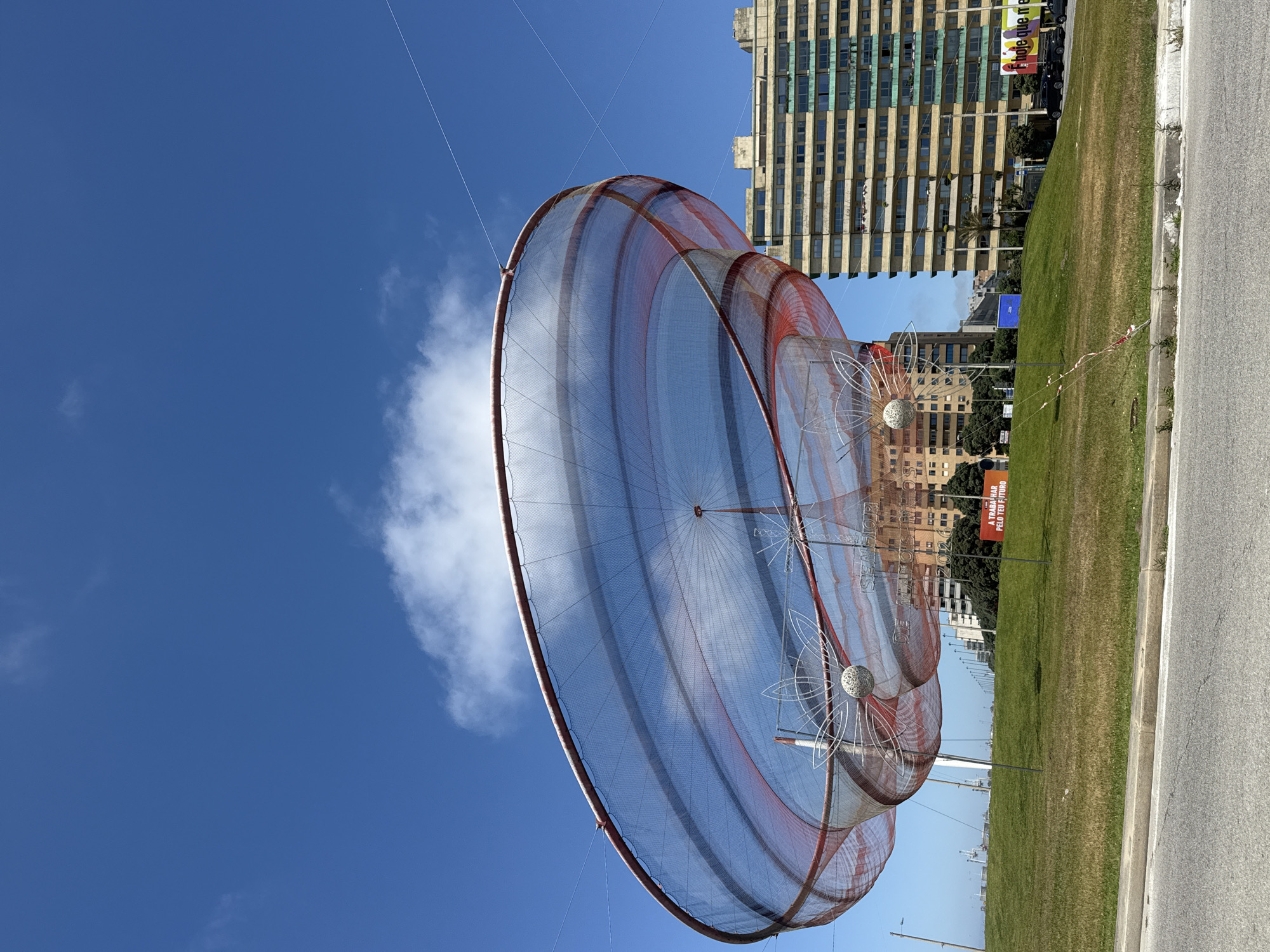 The image shows a public art installation located in Matosinhos, Portugal. The installation appears to be a large, circular structure made of netting, supported by cables. The structure features decorative, flower-like elements. The background includes an older high-rise building with multiple windows and balconies, a grassy median, a road, and a clear blue sky with a few clouds. A red sign displaying Portuguese text is also visible. The lighting indicates a sunny day.