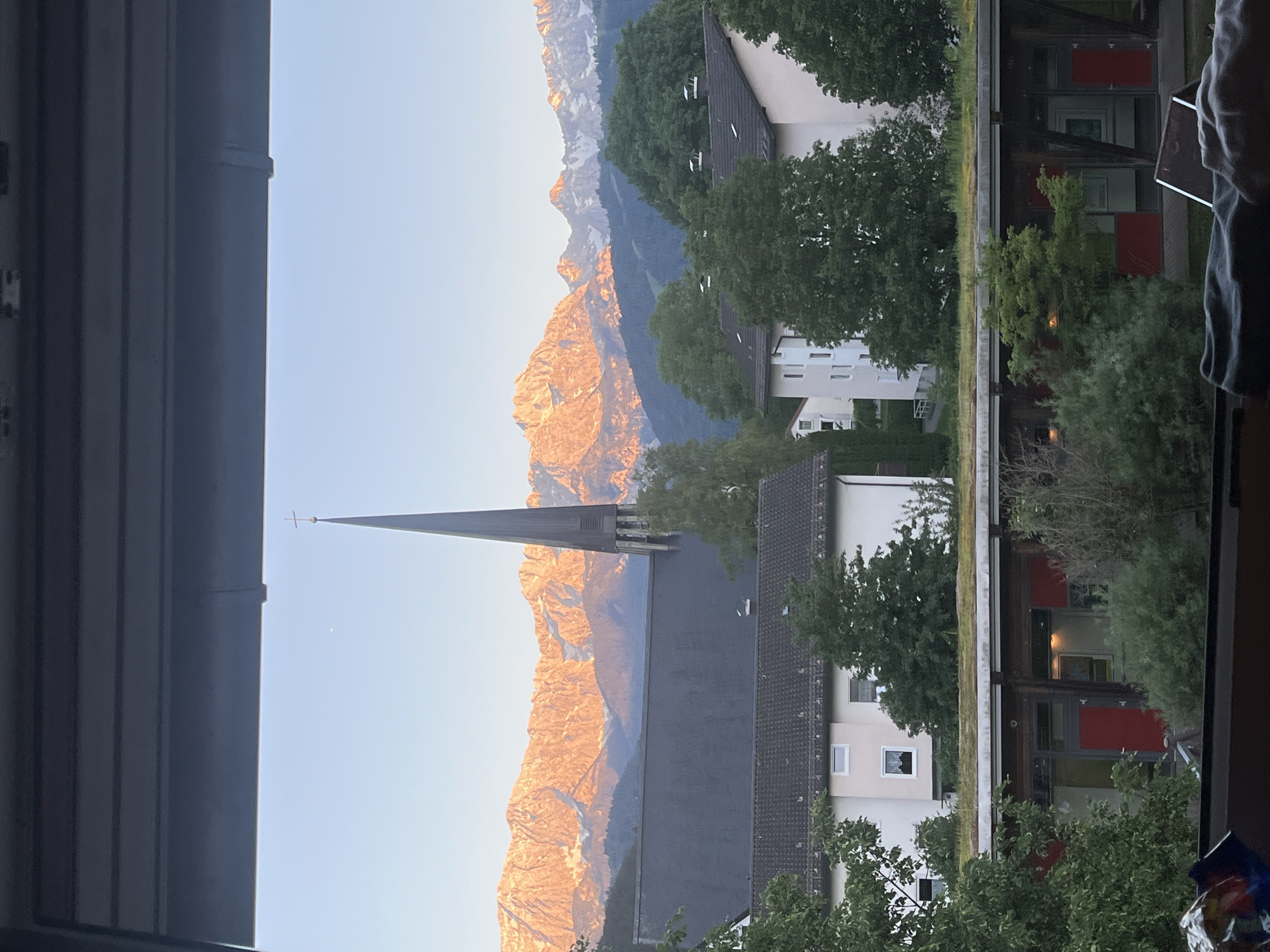 The image shows a view from a window overlooking a church steeple and the mountains beyond, likely in Garmisch-Partenkirchen, Germany. The church is a prominent feature, with a tall, pointed steeple topped by a cross. In the background, mountains stretch across the horizon, bathed in golden sunlight. Residential buildings are visible, interspersed with trees. The photo is taken through a window, with the frame and blinds partially visible on the left and some interior details on the right.