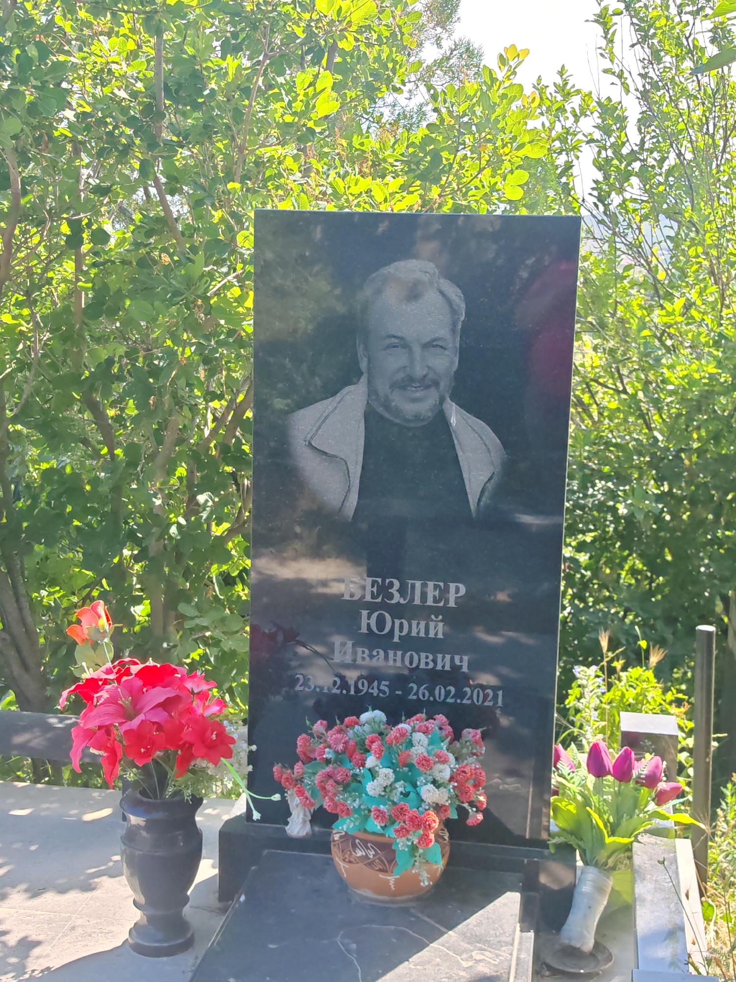 The image depicts a headstone in a cemetery, set against a backdrop of green foliage under a bright sky. The headstone is made of dark, polished stone and features a portrait of a man etched into its surface. Below the portrait, text is engraved, appearing to be a name and dates. Specifically, the text reads "ВЕЗЛЕР ЮРИЙ ИВАНОВИЧ 23.12.1945 - 26.02.2021". In front of the headstone, there are several bouquets of artificial flowers. To the left, a dark vase holds a large arrangement of red and pink flowers. To the right of the headstone, a brown decorative pot contains a bouquet of pink and white flowers, and further to the right, a white vase holds a bunch of purple tulips. The ground in front of the headstone appears to be paved with stone slabs. The overall scene is a typical memorial setting. The location is specified as the city of Supsekh, Russia.