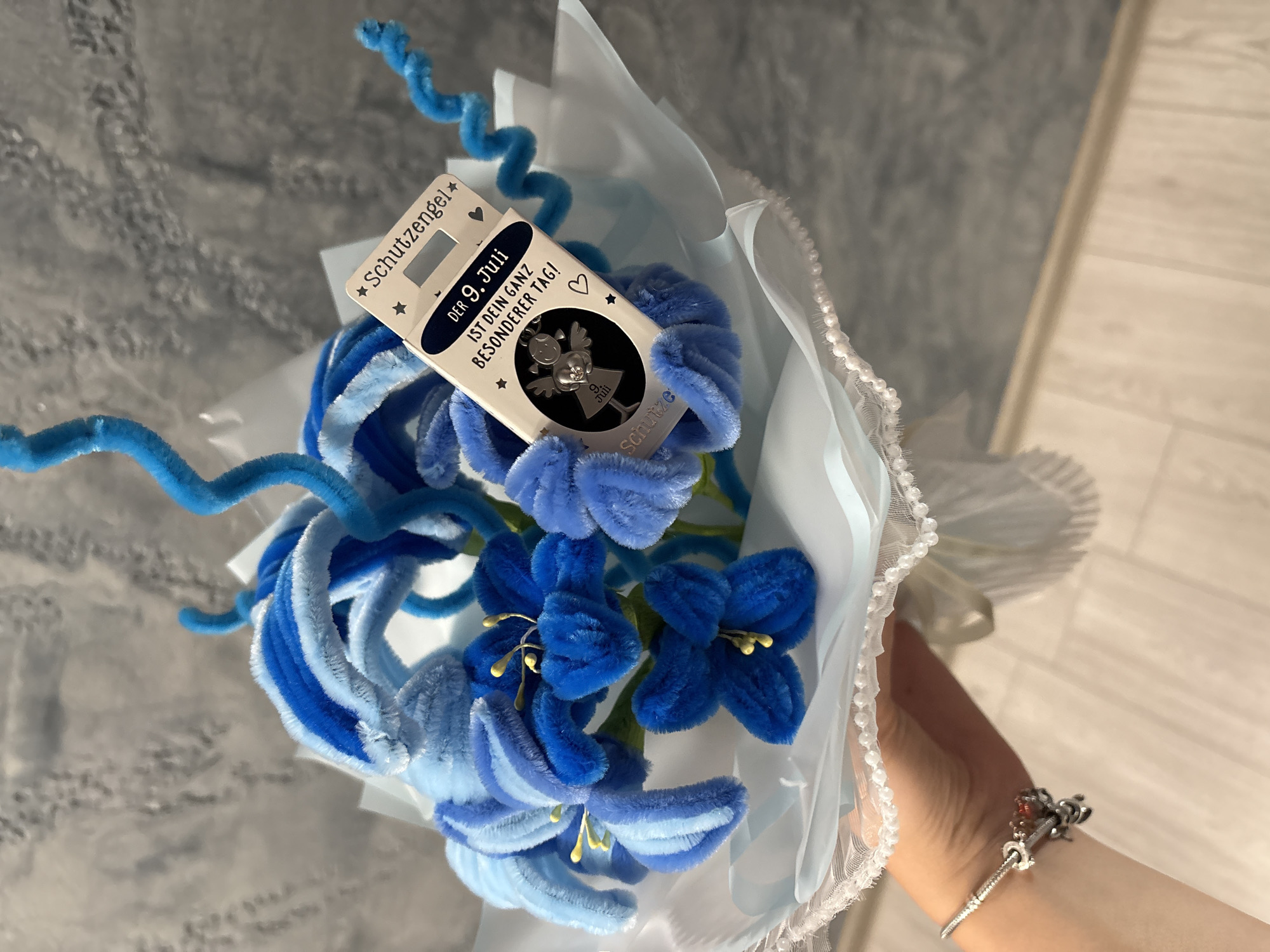 A close-up, eye-level shot shows a bouquet of blue pipe cleaner flowers. Attached to the bouquet is a small silver charm in a clear plastic package with a white card that reads "Schutzengel" and "DER 9. JULI IST DEIN GANZ BESONDERER TAG!". A hand with a silver charm bracelet is visible holding the bouquet from below. The background is a textured gray wall. The image is not relevant to Panagyurishte, Bulgaria.