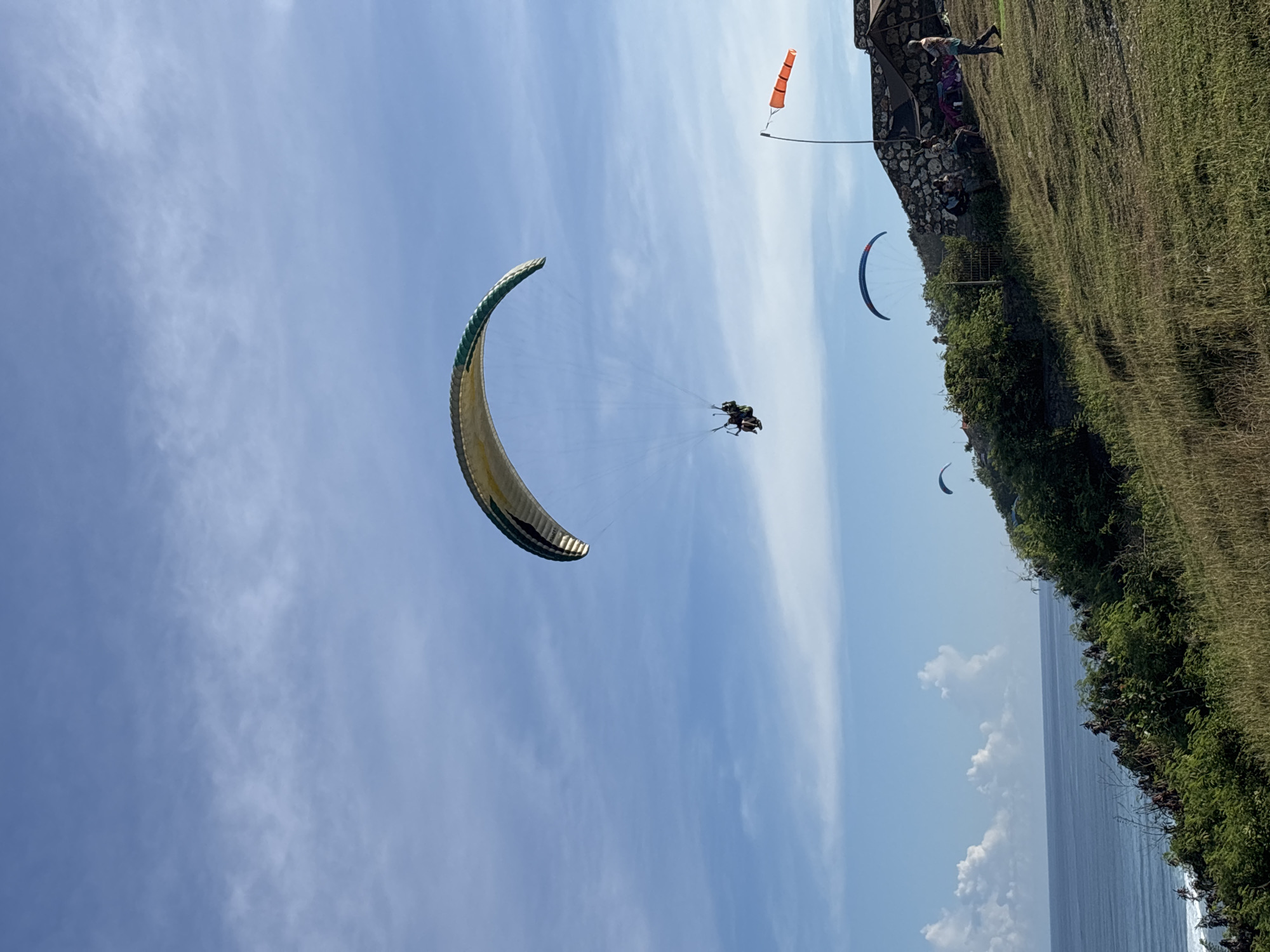 The image shows paragliders in the sky over a coastal area. The primary paraglider, yellow and green, is carrying two people and located in the left half of the frame against a blue sky with scattered clouds. In the background on the right side, there's a coastal cliff with green vegetation and a small structure, possibly a building or a viewing point. A windsock is visible near the cliff. There are other paragliders in the far distance closer to the cliffs. Based on the location, this is likely in Kuta Selatan, Indonesia, known for its coastal scenery and paragliding activities. The photograph captures the sport of paragliding set against a tropical coastal backdrop.