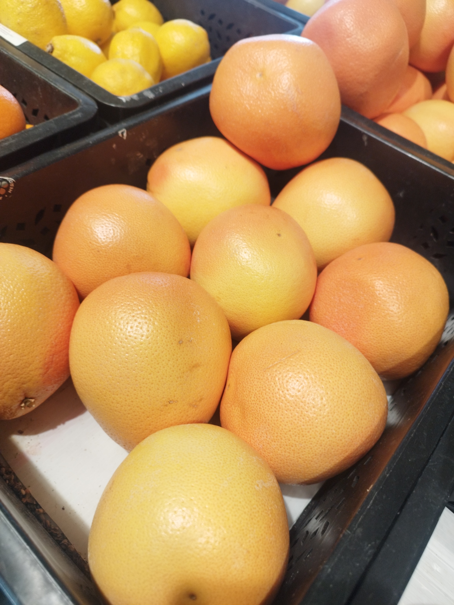 The image displays a close-up, overhead view of a bin of grapefruits and lemons. The grapefruits are in the foreground, with their textured orange rinds filling the majority of the frame. They are arranged in a dense cluster, some overlapping. In the background, to the left, a bin of bright yellow lemons is visible. The lighting is bright, highlighting the fruits' textures and colors. The scene appears to be in a market or grocery store setting, given the display bins. No specific location in Balashikha, Russia, can be identified from the image. There are no people or actions depicted. The environment is limited to the fruit bin and its contents.