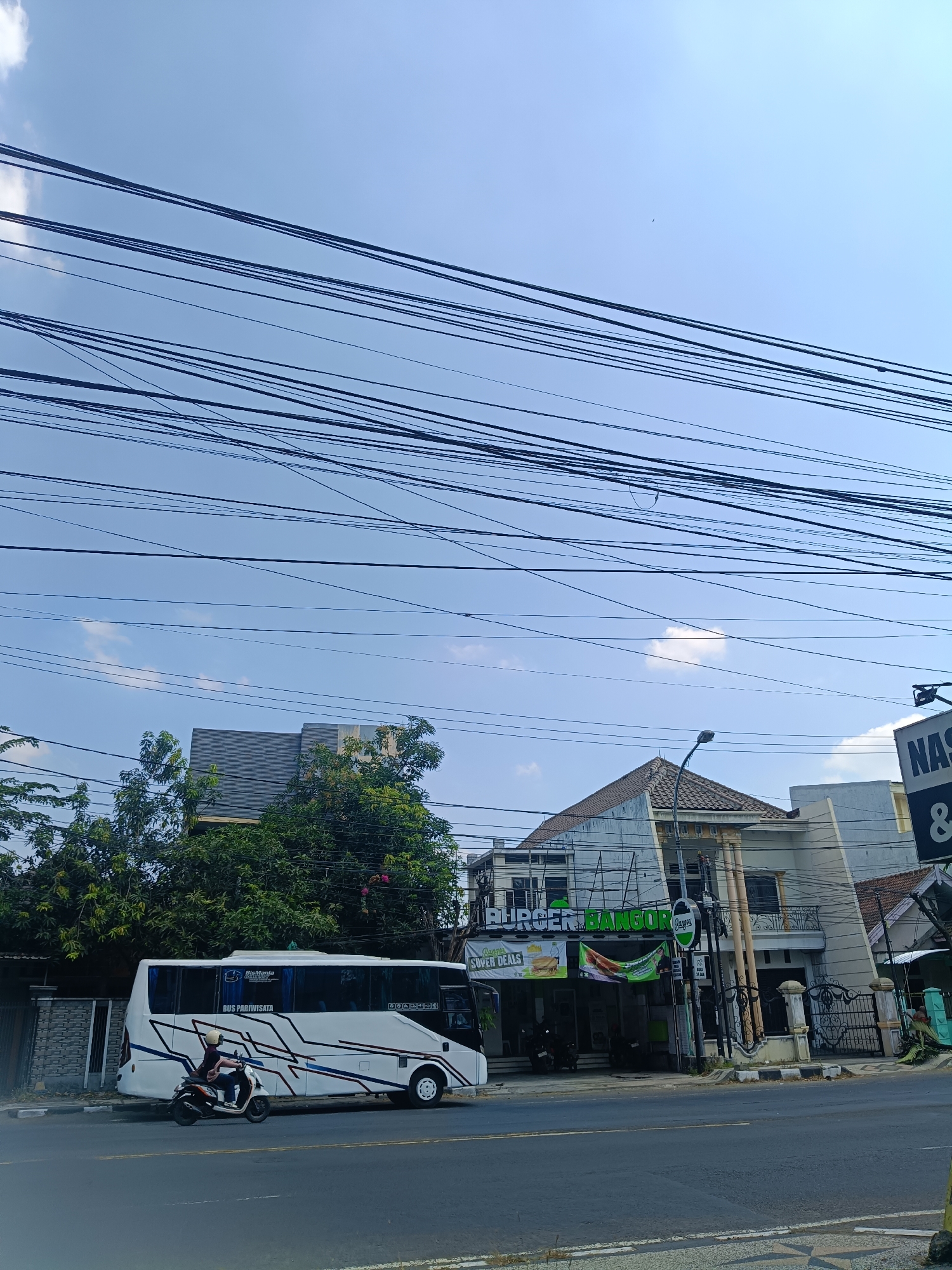 In Dapur, Indonesia, on a clear day, a white passenger bus with the text "BUS PARIWISATA" is parked alongside a road. A person on a scooter rides past the bus. In the background, a building with a sign for "BURGER BANGOR" is visible, along with other structures and abundant overhead power lines crisscrossing the sky.