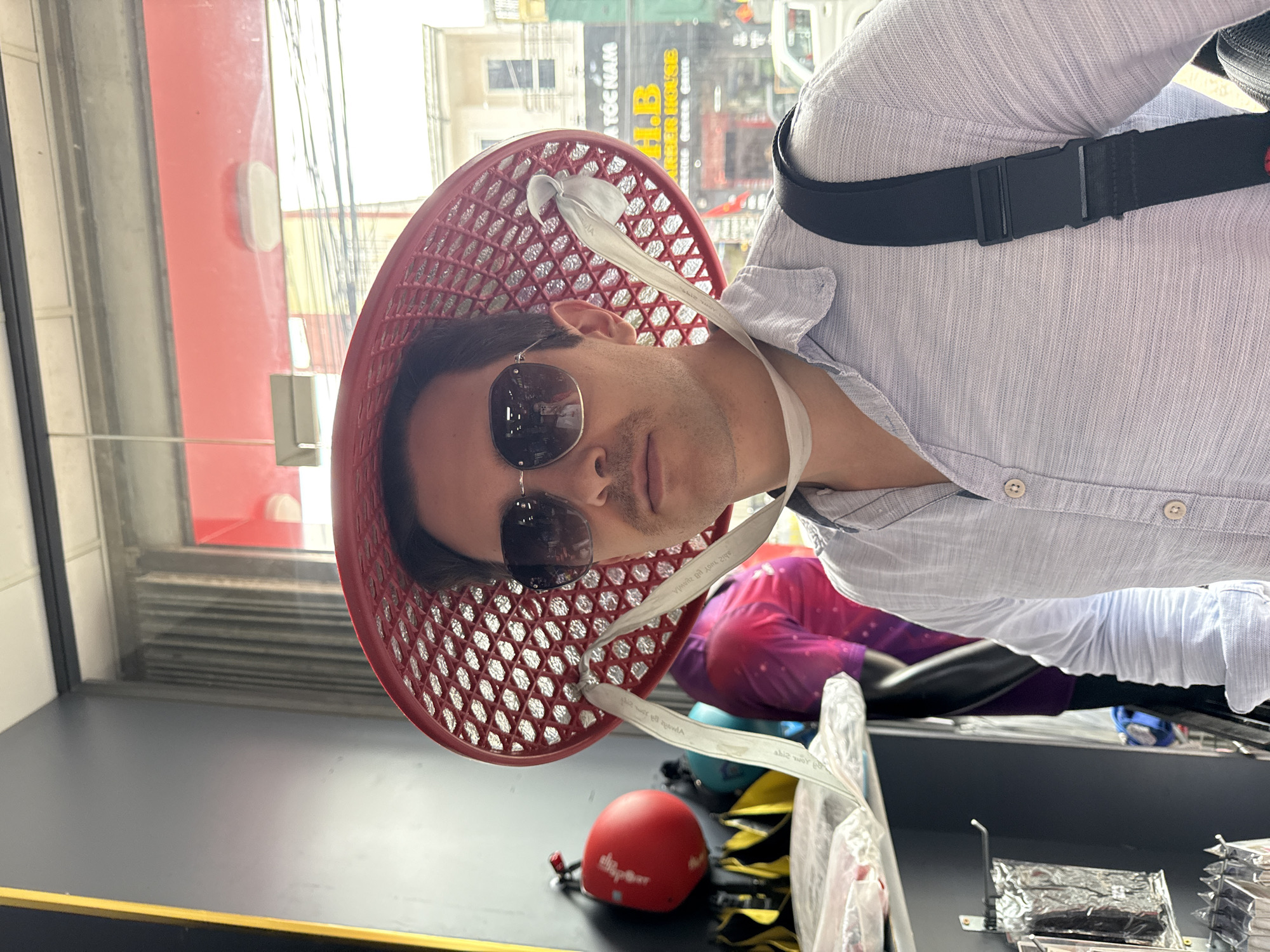 A man wears a traditional Vietnamese conical hat made from red plastic mesh, secured with white fabric straps. He is wearing sunglasses and a light blue button-down shirt. He has a black strap across his body. The background shows a storefront window with reflections of buildings and signs in the city of Da Lat, Vietnam. Below him on the counter is a red helmet.