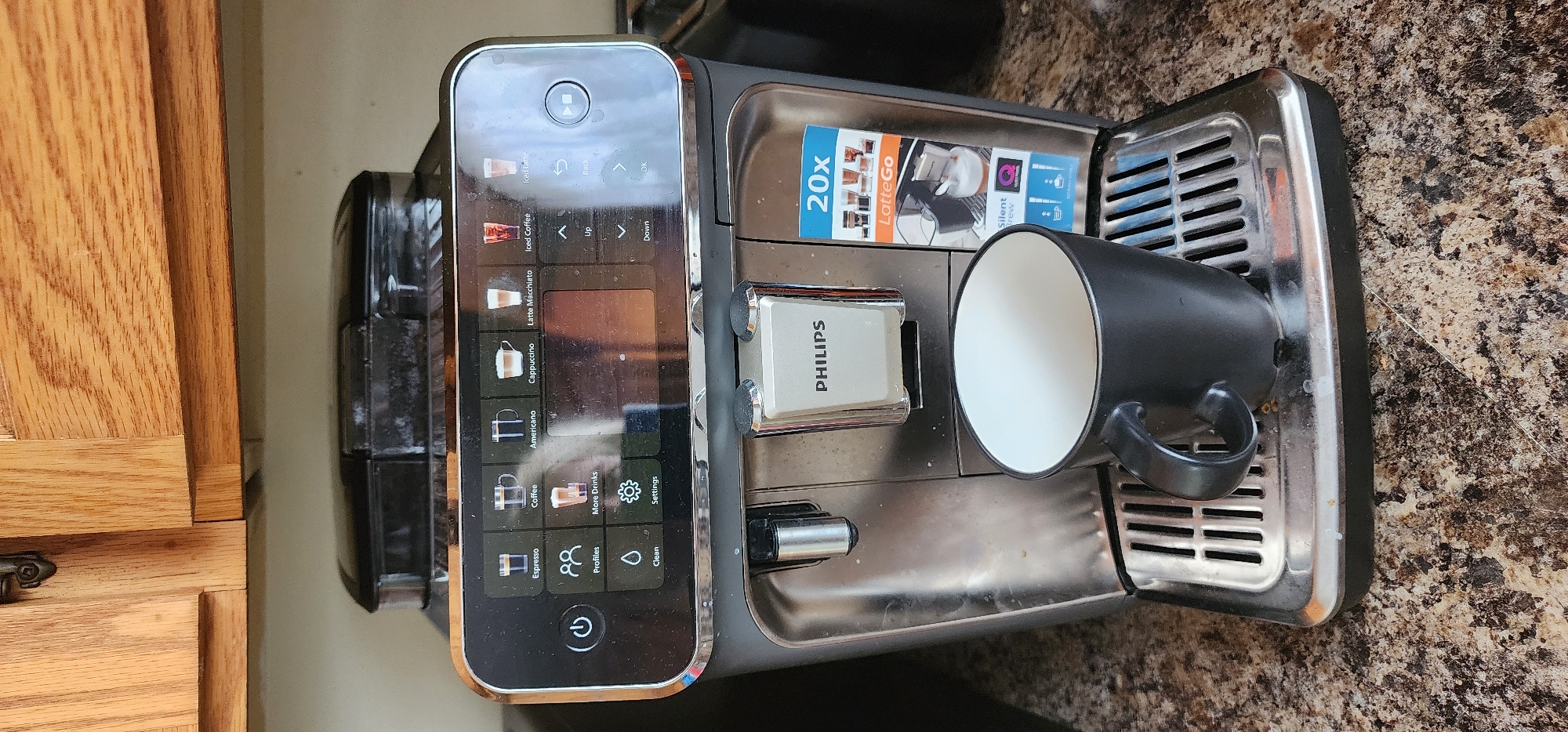 This is a head-on, medium shot of a Philips espresso machine, positioned on a speckled brown countertop. Above the machine, wooden kitchen cabinets are visible. The espresso machine features a digital display screen showing various coffee options and settings. Below the screen, the Philips logo is displayed on a chrome panel. A black mug sits on the drip tray of the machine, ready to receive coffee. A small, blue and white advertisement for "LatteGo" is affixed to the right side of the machine. There are no people present in the image. The scene is indoors, likely in a kitchen setting. The precise location in St. John's, Canada, cannot be determined from the image alone.