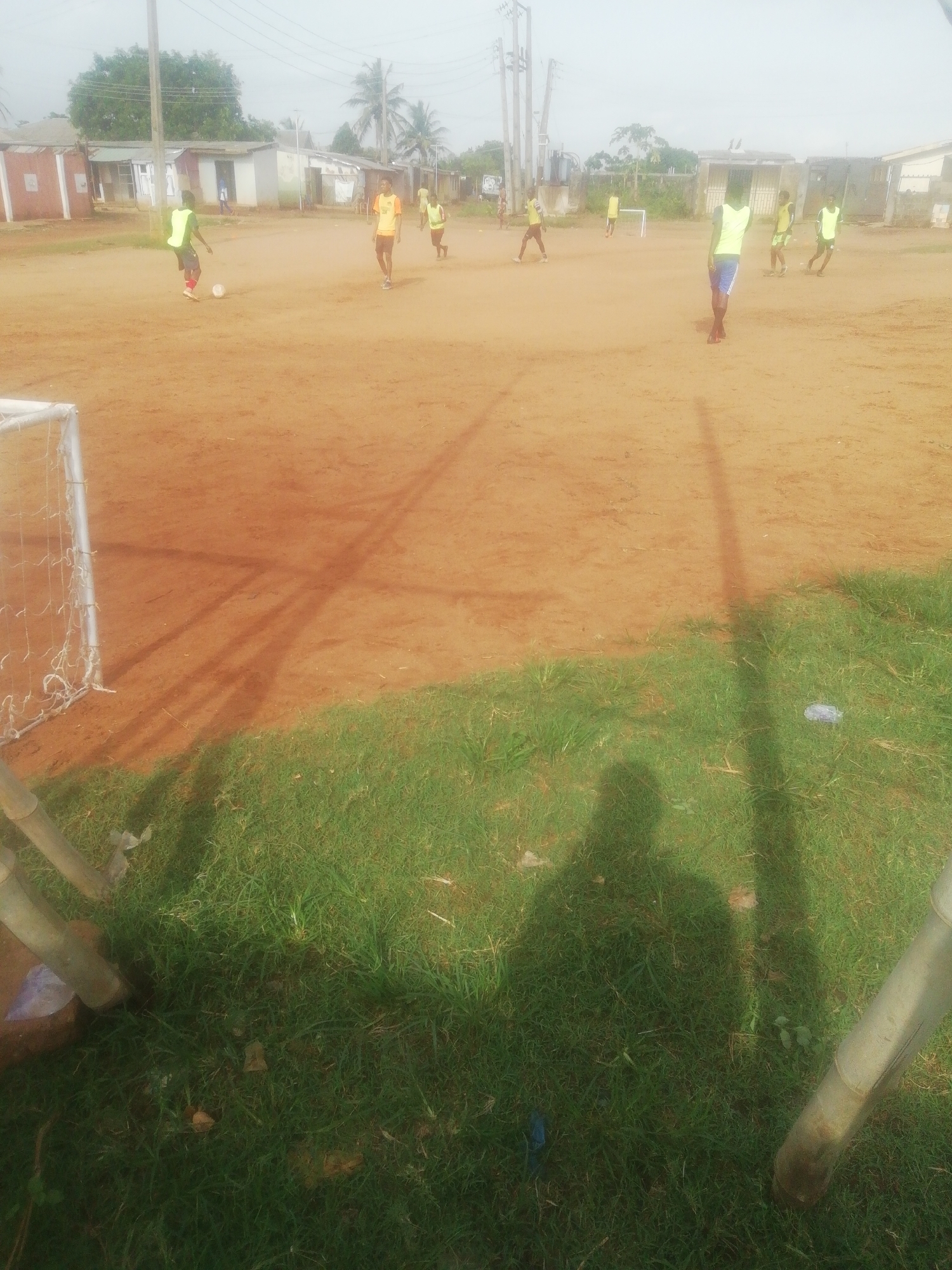 The image shows a group of young men playing soccer on a dirt field in Ikorodu, Nigeria. The players are wearing vests, and a soccer ball is visible near one player's feet. A soccer goal is present on the edge of the field. Houses and power lines can be seen in the background. The foreground contains a patch of green grass and the shadow of the photographer.