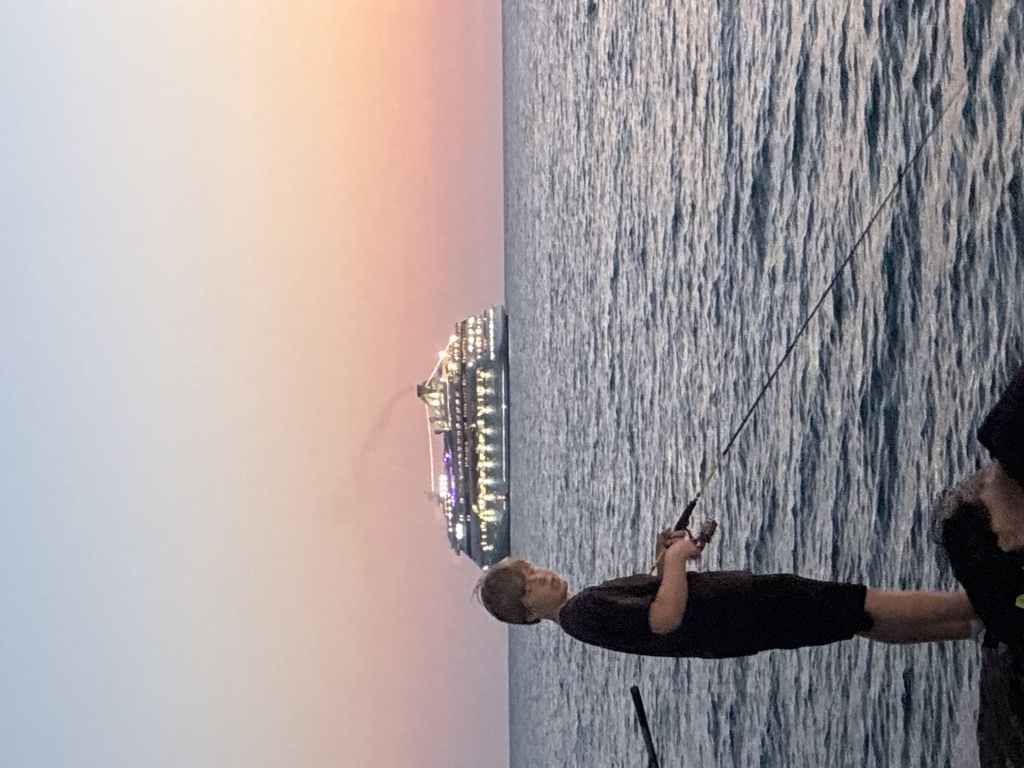 A young boy stands on a rocky shore, fishing in the sea. He is wearing a dark t-shirt and shorts. His fishing rod is extended towards the water. In the background, a large cruise ship is visible on the horizon, its lights illuminated against the twilight sky. The sky displays a gradient of pale blue at the top, transitioning to soft pink and orange hues near the horizon. The sea is calm with gentle ripples. The scene appears to be set at dusk or dawn, likely near the coast of Sochi, Russia, given the presence of a cruise ship and the serene seascape often found in that region.