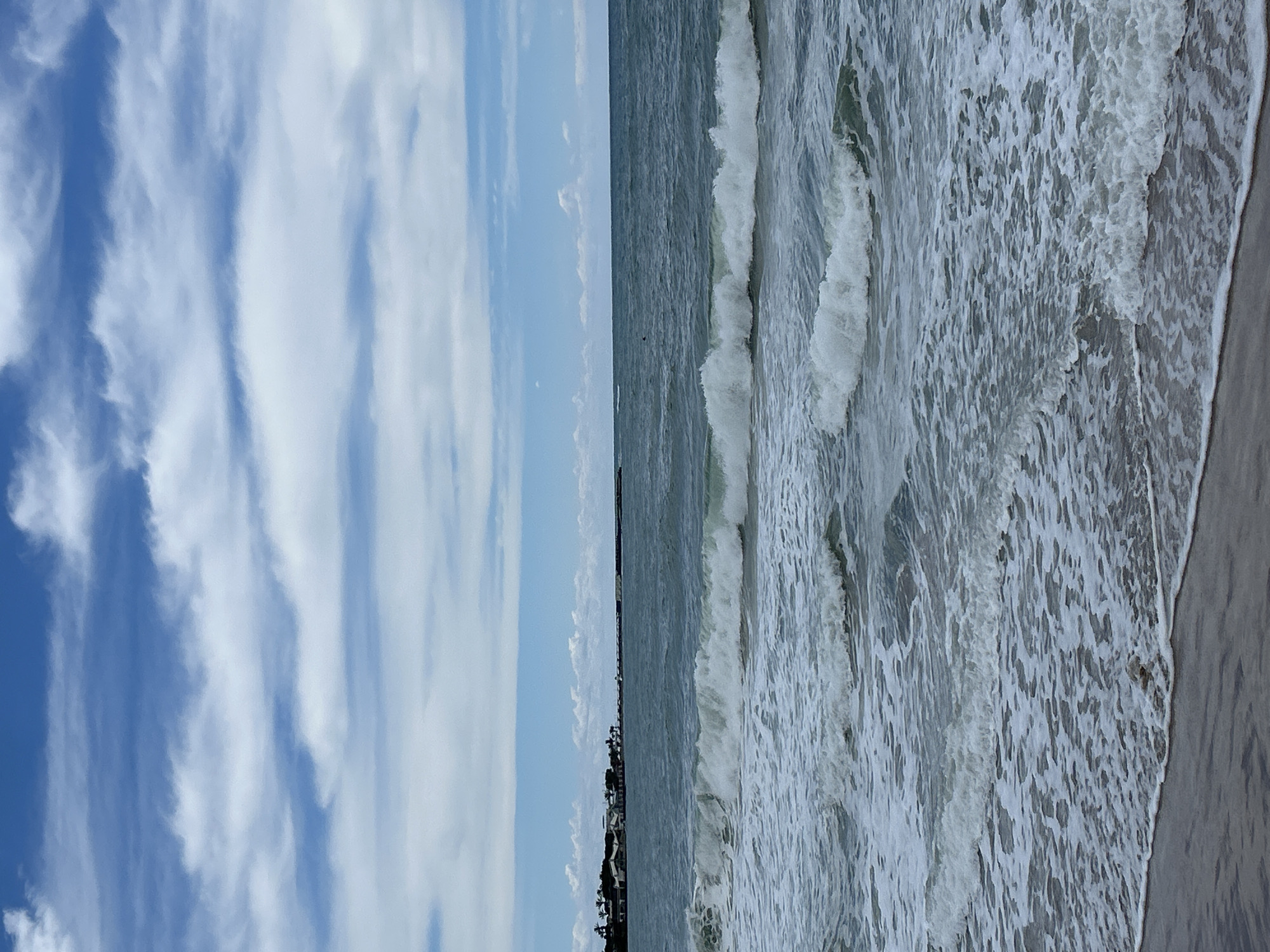A landscape-oriented photograph captures a shoreline scene, likely taken on a beach, with a clear delineation between sky, sea, and sand. The image appears to be rotated.
On the left side of the frame, the sky dominates, featuring a vibrant blue hue interspersed with elongated, wispy white clouds that suggest altitude and movement. These clouds exhibit a striated pattern, adding texture to the aerial expanse.
Adjacent to the sky, the sea stretches out, with visible waves breaking and rolling towards the shore. The water is a mix of blues and grays, indicative of depth and potential turbidity. The foamy white caps of the waves contrast sharply with the darker water beneath, enhancing the sense of motion.
The shoreline is depicted with a sandy beach in the foreground, transitioning from wet sand closest to the water to drier sand further inland. The color of the sand is a neutral beige, providing a grounding element to the composition.
In the far distance, structures resembling houses or buildings are barely visible along the horizon, suggesting a coastal development or urban area nearby.
The scene captures the dynamic interplay between the elements—sky, water, and land—with the suggestion of human presence in the distance. Without further evidence, it is difficult to verify that the picture was taken in Alanya, Turkey.