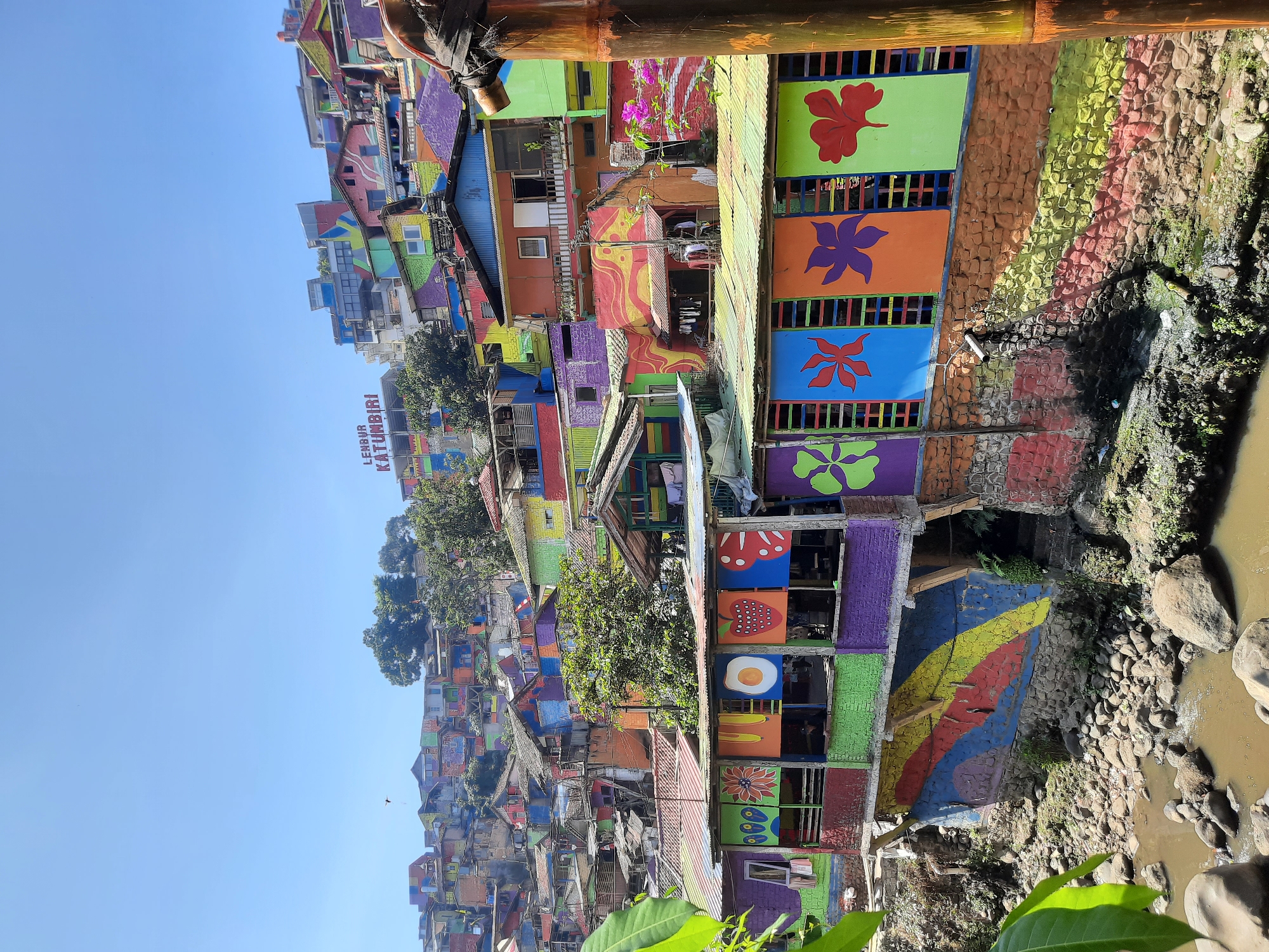 This is a photograph of Kampung Pelangi, a village in Bandung City, Indonesia, transformed into a vibrant tourist attraction through colorful renovations. The image captures a hillside densely populated with houses, all painted in a rainbow of bright colors, including pink, orange, yellow, green, blue, and purple. The houses are built close together, with many multi-story structures.
In the foreground, a section of the village features shops or stalls with corrugated metal roofs. The fronts of these stalls are adorned with colorful painted panels depicting various motifs, such as flowers, a fried egg, and strawberries. Below these stalls, the walls are painted with bold, abstract colorful stripes. The ground level transitions into a rocky riverbed with murky water flowing.
Further up the hill, the colorful houses continue to climb, creating a visually striking landscape. Trees are interspersed among the buildings, and the sky above is a clear, bright blue. A sign that reads "LENYUR KATUMBARI" is visible on a building higher up the hillside. The overall impression is one of a densely packed, lively community that has embraced a highly artistic and colorful aesthetic. There are no visible people prominently featured in the image. The lighting suggests it is daytime, with the sun casting shadows on the buildings and the ground.