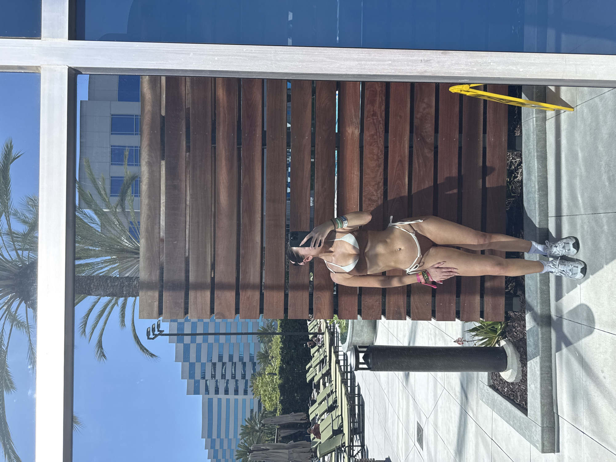 A woman in a white bikini is reclining on a wooden slat bench in an outdoor area. She is wearing white socks and gray sneakers. She is holding a phone, presumably taking a selfie.
The location appears to be a poolside or patio area in Irvine, California. The reflection in the glass shows a tall office building. A yellow "Wet Floor" sign is visible near the edge of the bench. A black bollard is situated nearby, with landscaping around its base. White tiles form the pavement. There are lounge chairs with green cushions. Palm trees are visible.