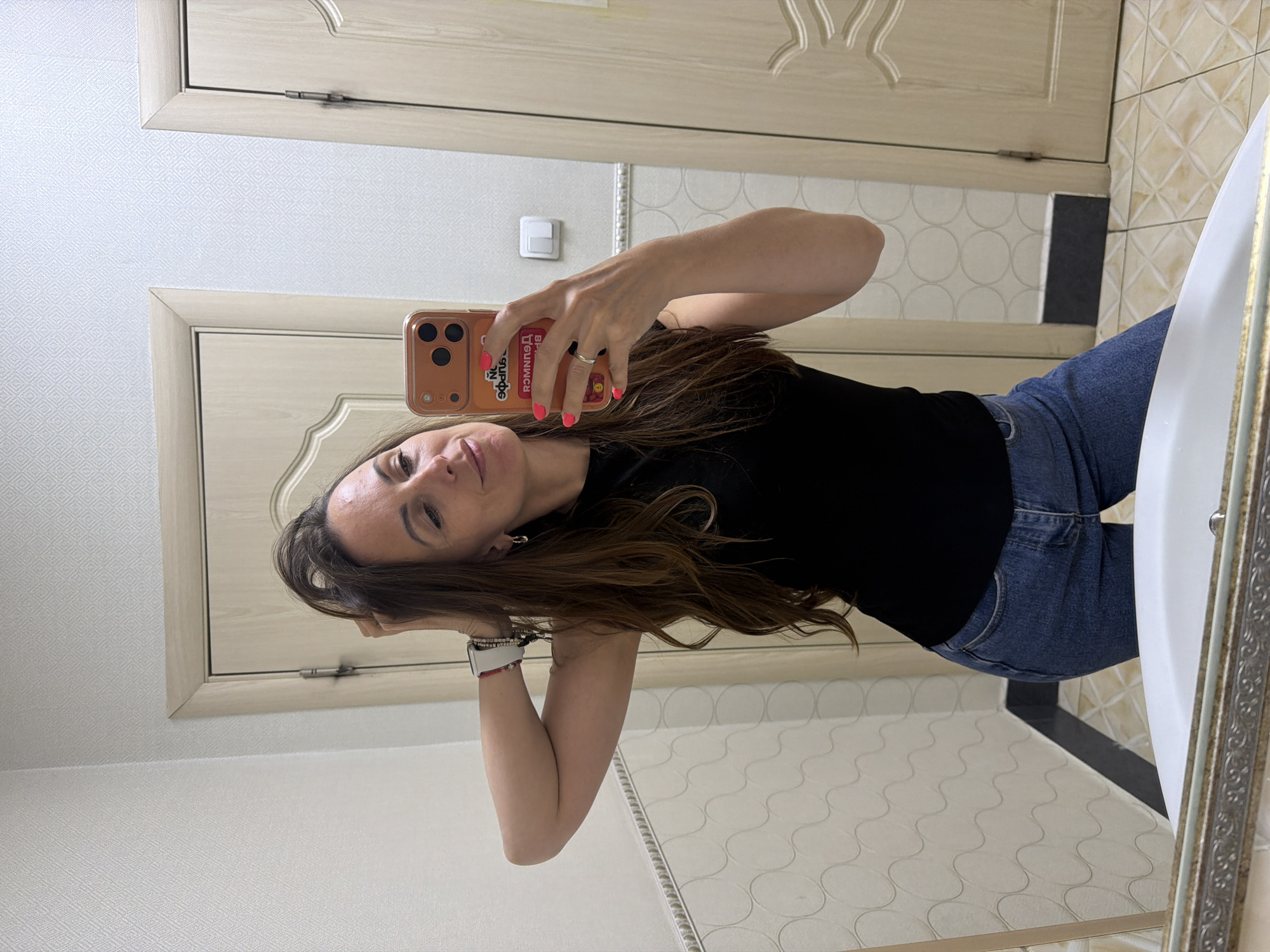 A woman is taking a selfie in a bathroom. She is wearing a black t-shirt and blue jeans. She is holding a phone with a cartoon-themed case in her right hand. Her hair is brown and wavy. The bathroom has beige and white patterned wallpaper and tile flooring. There is a white sink with a gold-framed mirror to the right. A beige door with decorative molding is visible in the background. The location is not identifiable as Birobidzhan, Russia.