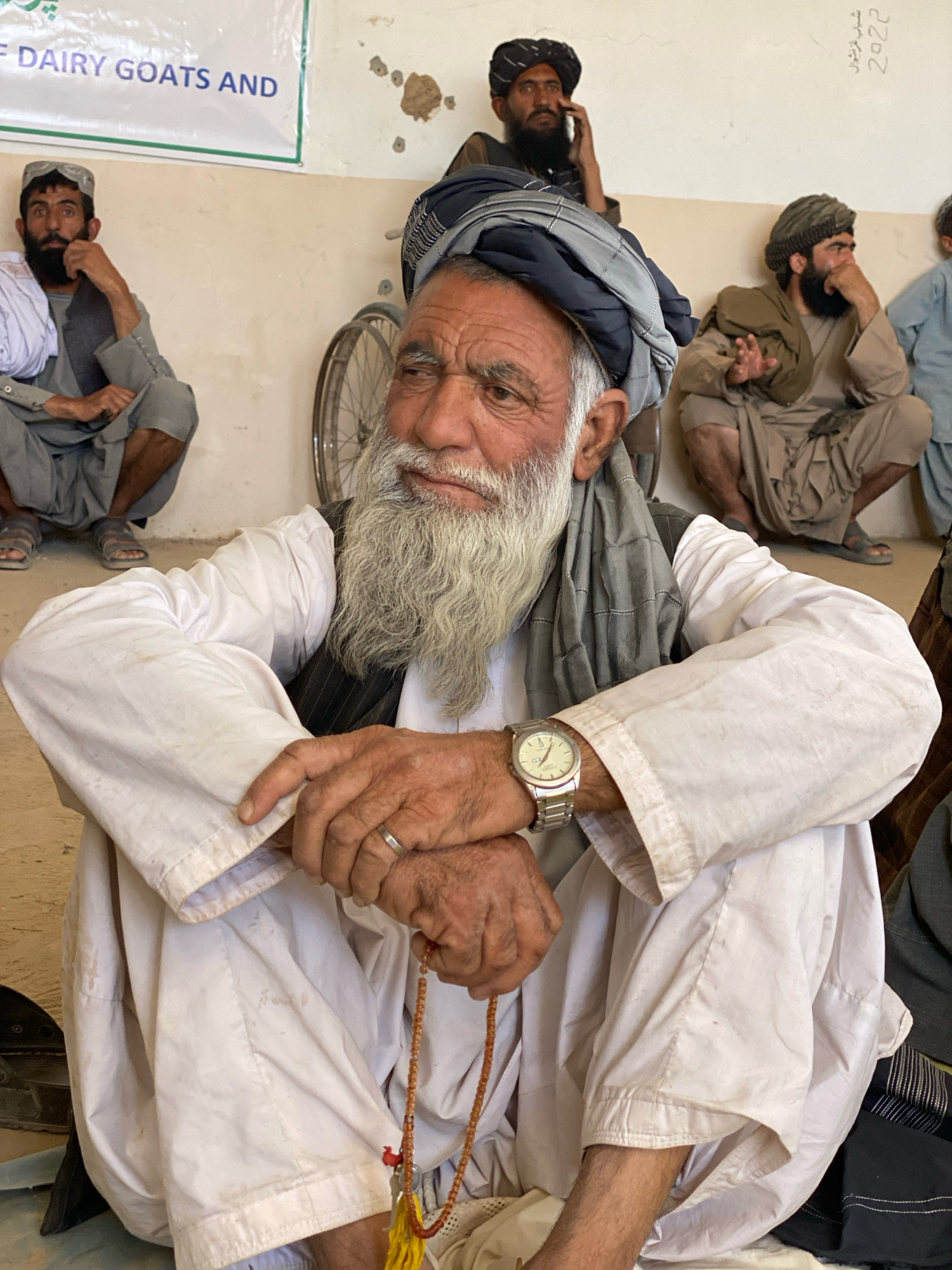 In Pasab, Afghanistan, a group of men are gathered indoors, possibly in a community center or a meeting hall, as indicated by the banner in the background that reads "DAIRY GOATS AND". The man in the foreground, an elder with a long white beard and a grey turban, is seated on the floor. He wears a white shalwar kameez and a dark vest. His left hand rests on his lap, and his right hand is clasped with a string of prayer beads. He wears a silver wristwatch on his left wrist and a ring on his right ring finger. Behind him, other men are seated in similar attire, some with turbans and traditional clothing. A bicycle wheel is visible in the middle ground, and the walls appear to be a light tan color with some rough patches. The overall lighting suggests an indoor setting during the daytime.