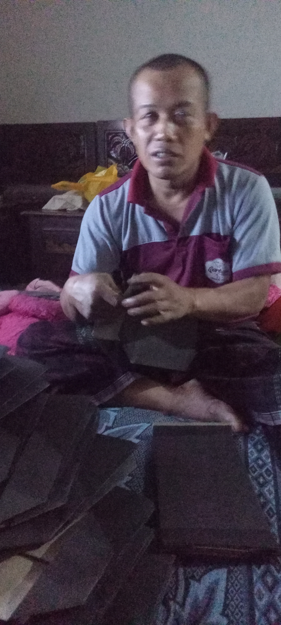 A man is sitting on a bed in a dimly lit room in Gempolklutuk, Indonesia, crafting what appear to be folded paper or cardboard objects. He is wearing a short-sleeved polo shirt with maroon and grey panels, and a maroon sarong is draped over his lap. His hands are engaged in manipulating a dark-colored object, likely part of the crafting process. Several similar folded objects are laid out in front of him on the bed and the floor. A wooden headboard is visible behind him, along with a yellow plastic bag on a dark surface. The room's ambiance is subdued due to the low lighting.