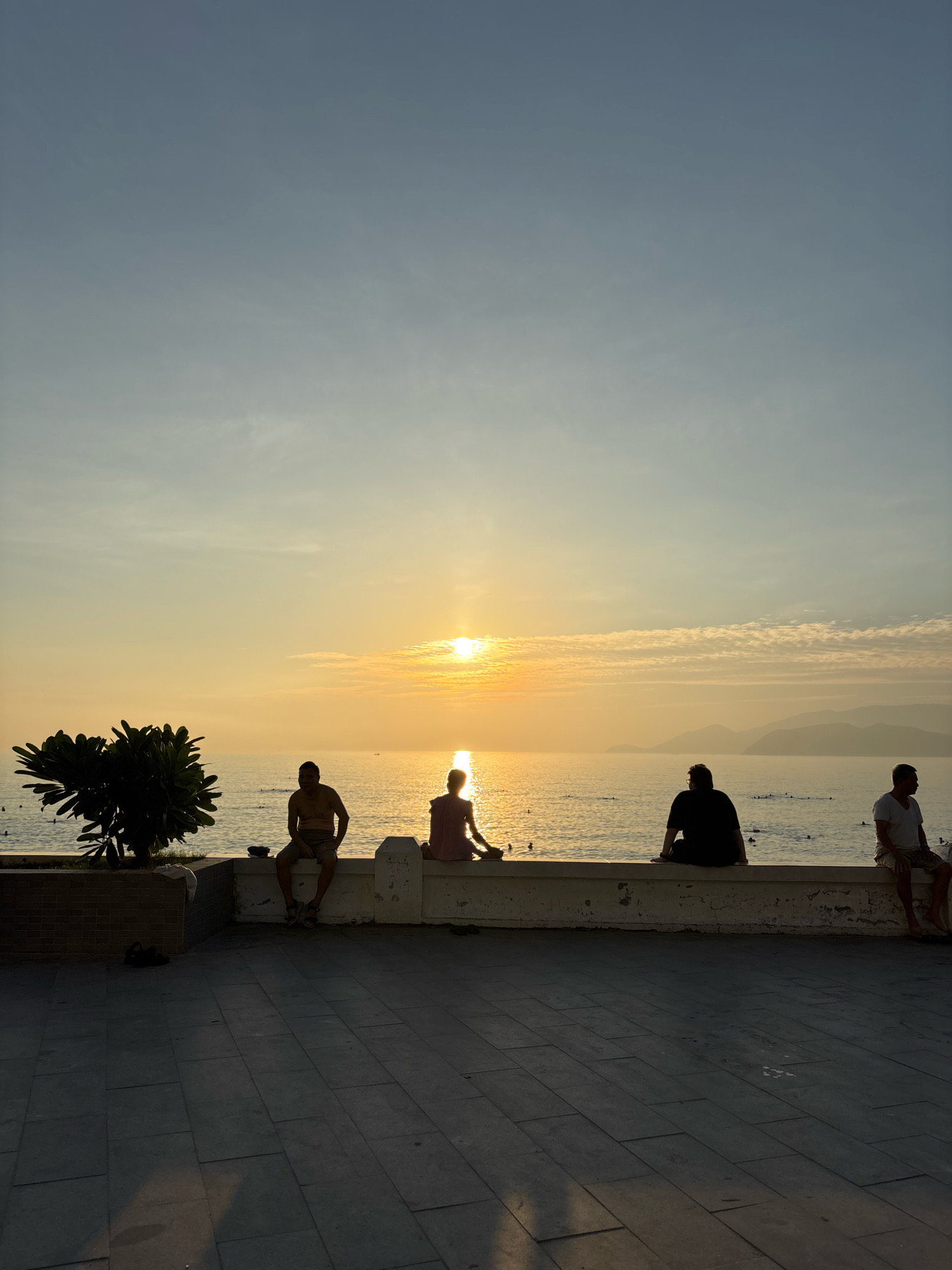 The image depicts a serene sunset scene along the coast of Nha Trang, Vietnam. In the foreground, a paved walkway with geometric tiling stretches across the bottom of the frame, casting long shadows from an unseen light source. A low, white concrete wall separates the walkway from the water, and atop this wall, five individuals are seated, silhouetted against the vibrant sky.
To the left, a dark-leaved tree with a thick trunk stands on the walkway, its branches reaching towards the sky. A small pile of dark sandals rests near its base.
The individuals are spaced along the wall, facing the ocean and the setting sun. From left to right, the first person is a man sitting with his legs extended. The second is a person in a light-colored top, seated with their legs crossed, facing the sun. The third is a man wearing a dark shirt, his back to the camera, seated with his legs drawn up. The fourth is a man in a light-colored t-shirt, his legs dangling over the wall, looking towards the ocean. The fifth person, partially visible on the far right, is also seated with their legs dangling.
The sea is calm, reflecting the warm golden hues of the setting sun. A few distant boats are visible on the water, and faint outlines of mountains can be seen on the horizon. Small figures are dispersed in the water, suggesting swimming or wading. The sky transitions from a pale blue at the top to a rich orange and yellow near the horizon, with wisps of clouds catching the last rays of sunlight. The overall atmosphere is tranquil and contemplative.