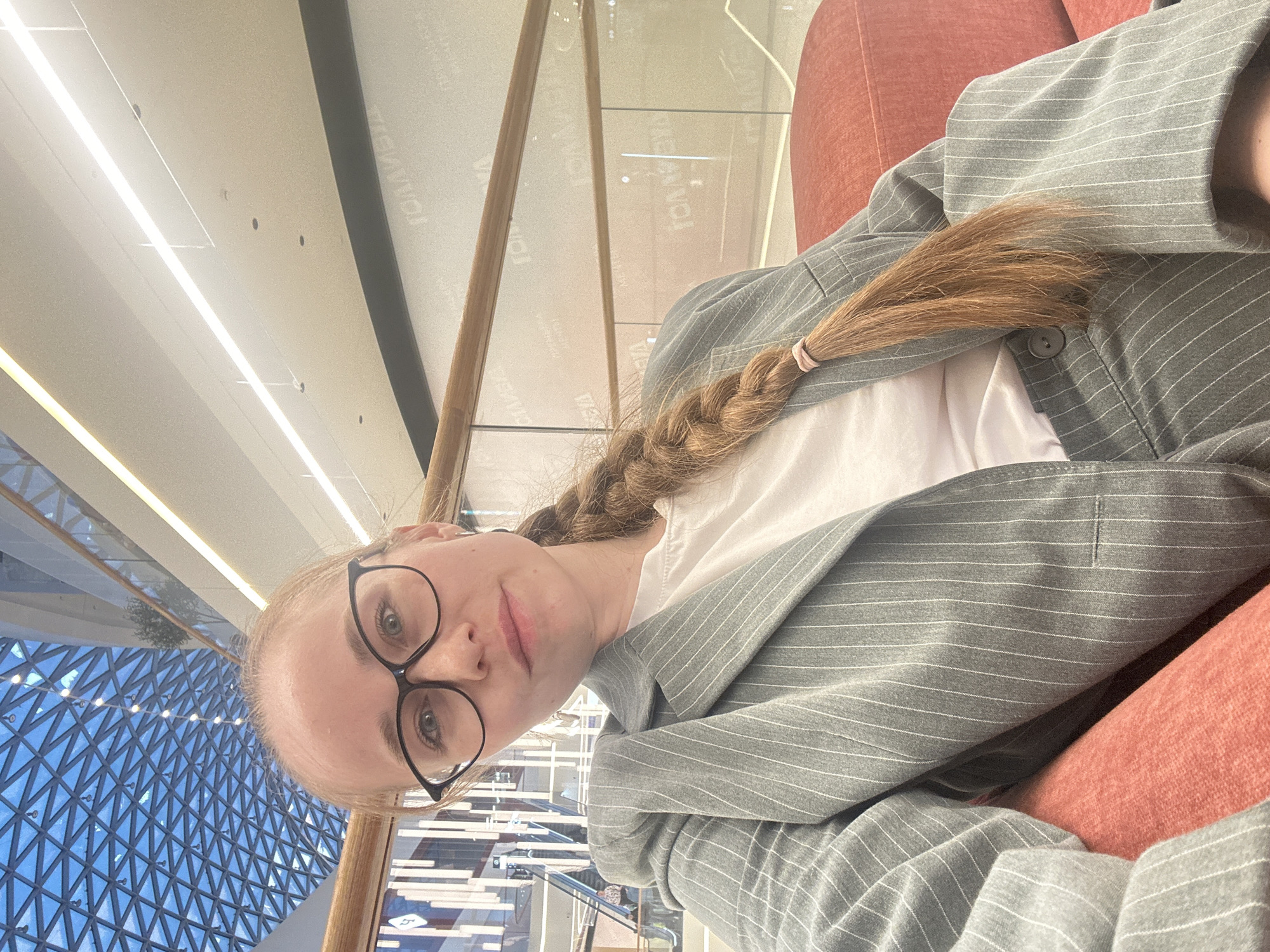 A woman with glasses is seated in a peach-colored chair and wearing a grey pinstripe blazer over a white shirt. Her blonde hair is braided. The location appears to be a modern building with a distinctive blue geometric structure visible through glass in the background. Based on the architectural style, specifically the modern, geometric facade, the building is likely located in Saint Petersburg, Russia, possibly the Lakhta Center or a similar contemporary structure.