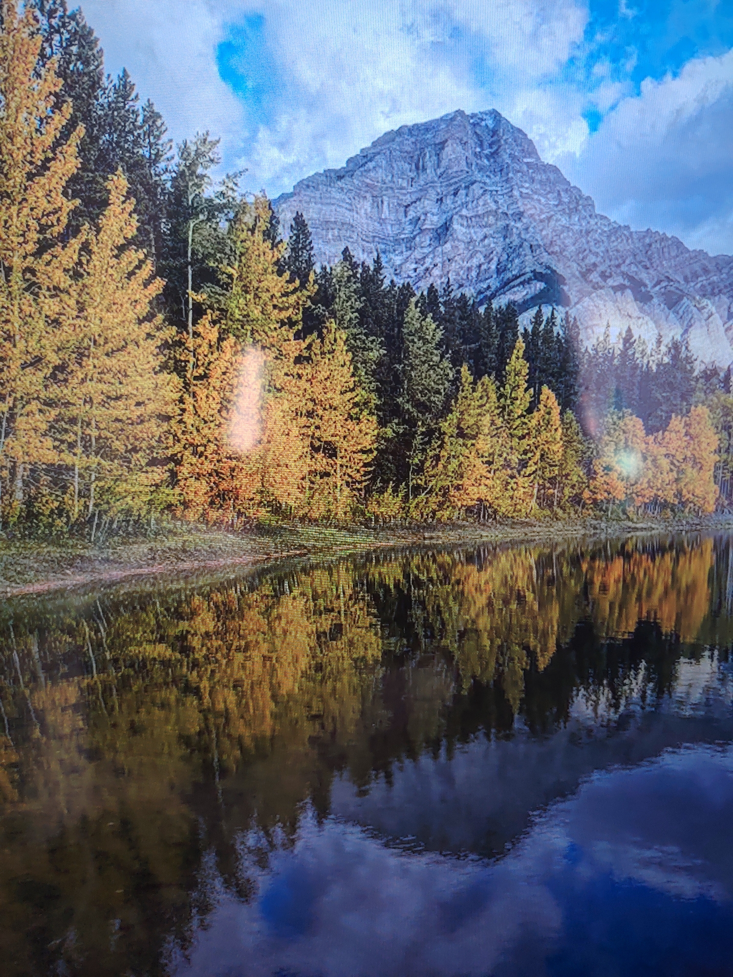 A mountain range towers over a placid body of water, with the reflection of the trees and sky visible in the water. The trees nearest to the water are primarily yellow and orange, suggesting it is autumn. The water is dark and has patches of blue where the sky is reflected. The mountain is rugged and gray with some snow. The sky is blue with scattered clouds. Location: Unknown, Unknown.